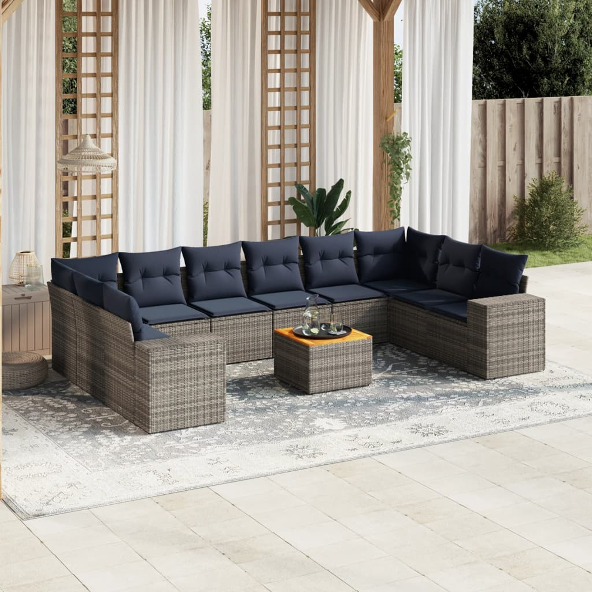Garten-Terrassenset mit grauem Korbgewebe, marinefarbenen Kissen und einem kleinen Holztisch auf einem gemusterten Teppich, unter einem weißen Vorhang.