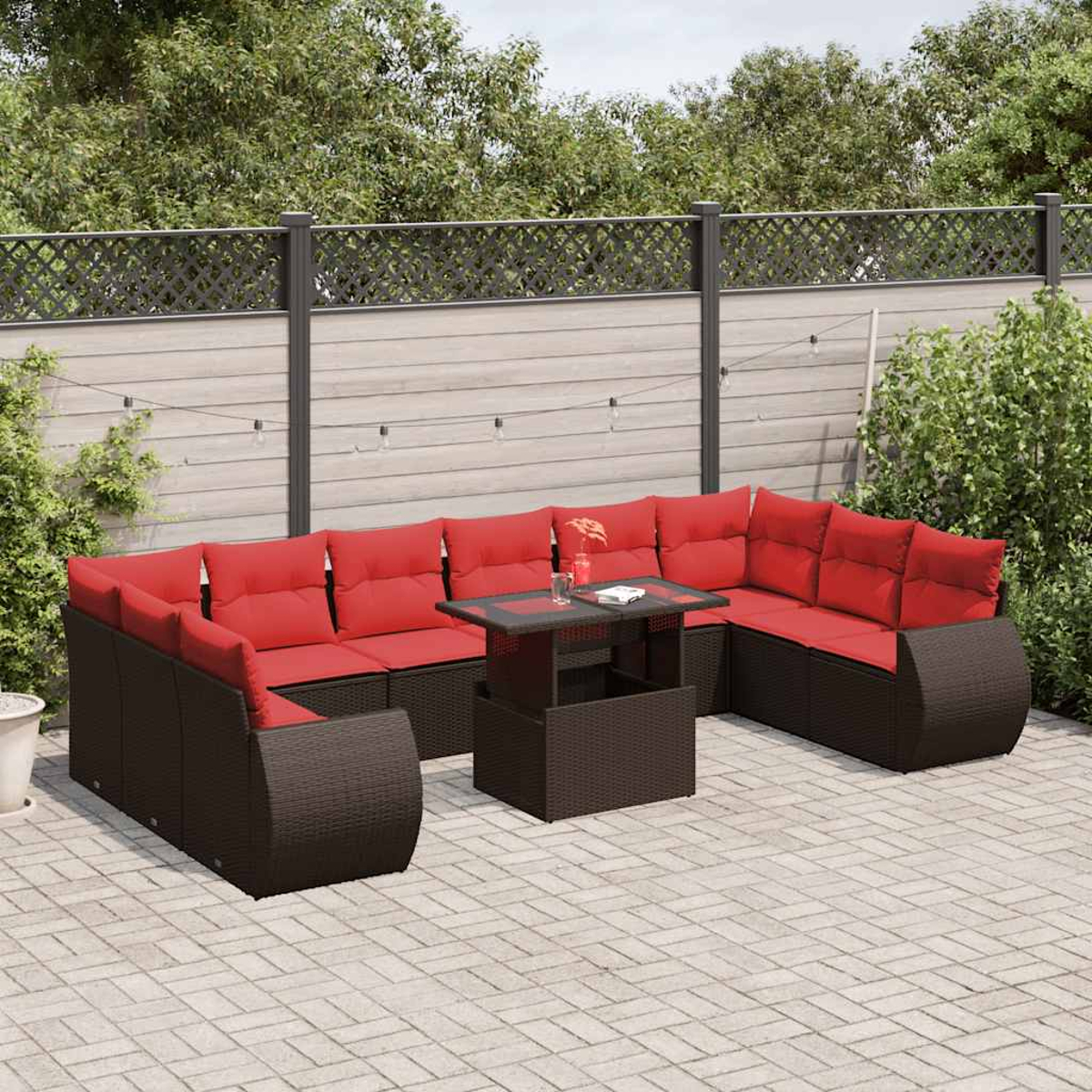 Garten-Terrassenset mit roten Kissen und braunen Korbflechtrahmen. Ein Tisch mit Glasplatte steht in der Mitte. Im Hintergrund sind Zaun und Bäume zu sehen.