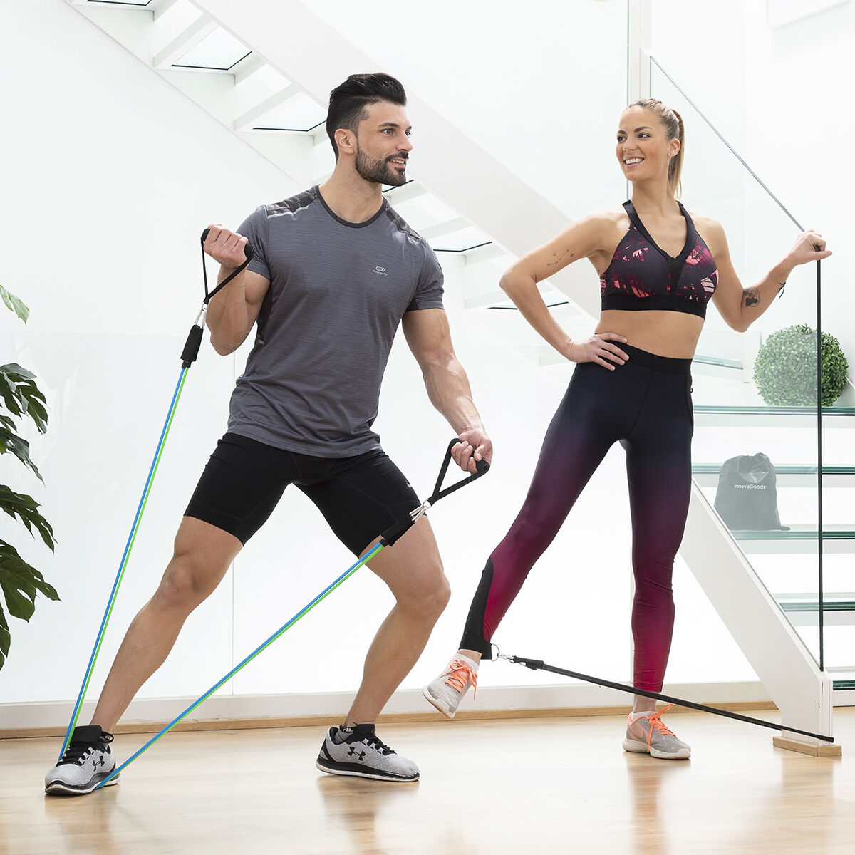 Ein Mann und eine Frau trainieren in einem hellen Raum mit Widerstandsbändern. Sie tragen Sportkleidung und lächeln beim Training.