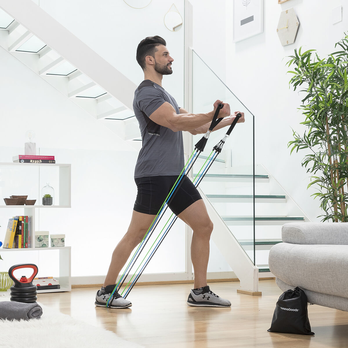 Mann trainiert mit Widerstandsbändern. Er befindet sich in einem hellen Raum mit Treppe, Couch und Fitnessgeräten im Vordergrund.