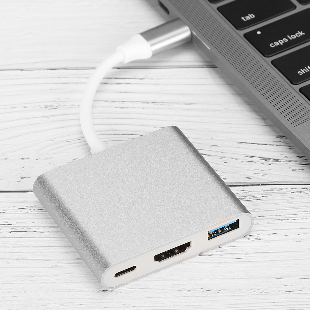 Ein silberner USB-C-Hub, der mit einem Laptop verbunden ist. Der Hub hat Anschlüsse für HDMI, USB und Aufladen. Der Hintergrund ist eine Holzoberfläche.