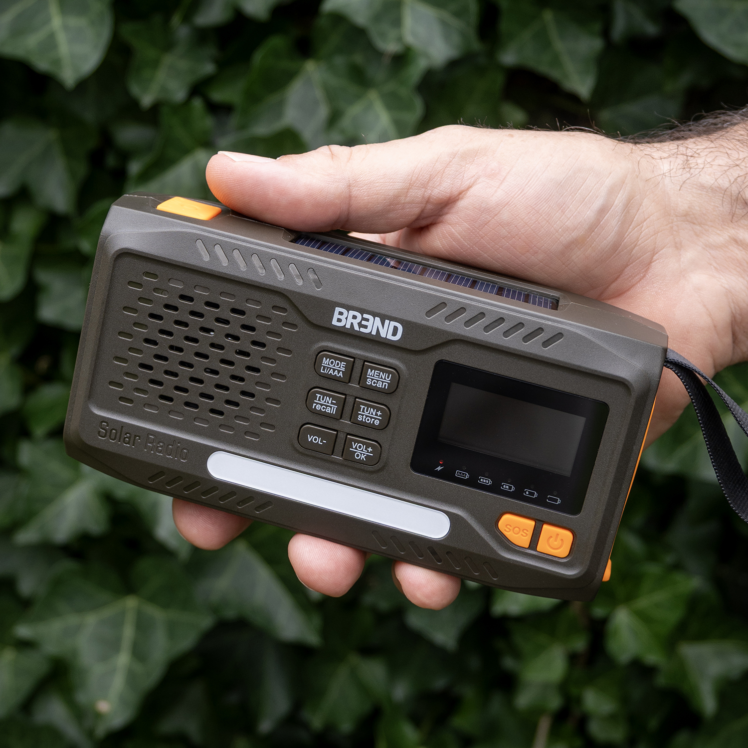 A hand holds a solar radio against a green leafy background. The radio is olive green with orange accents and a small display.