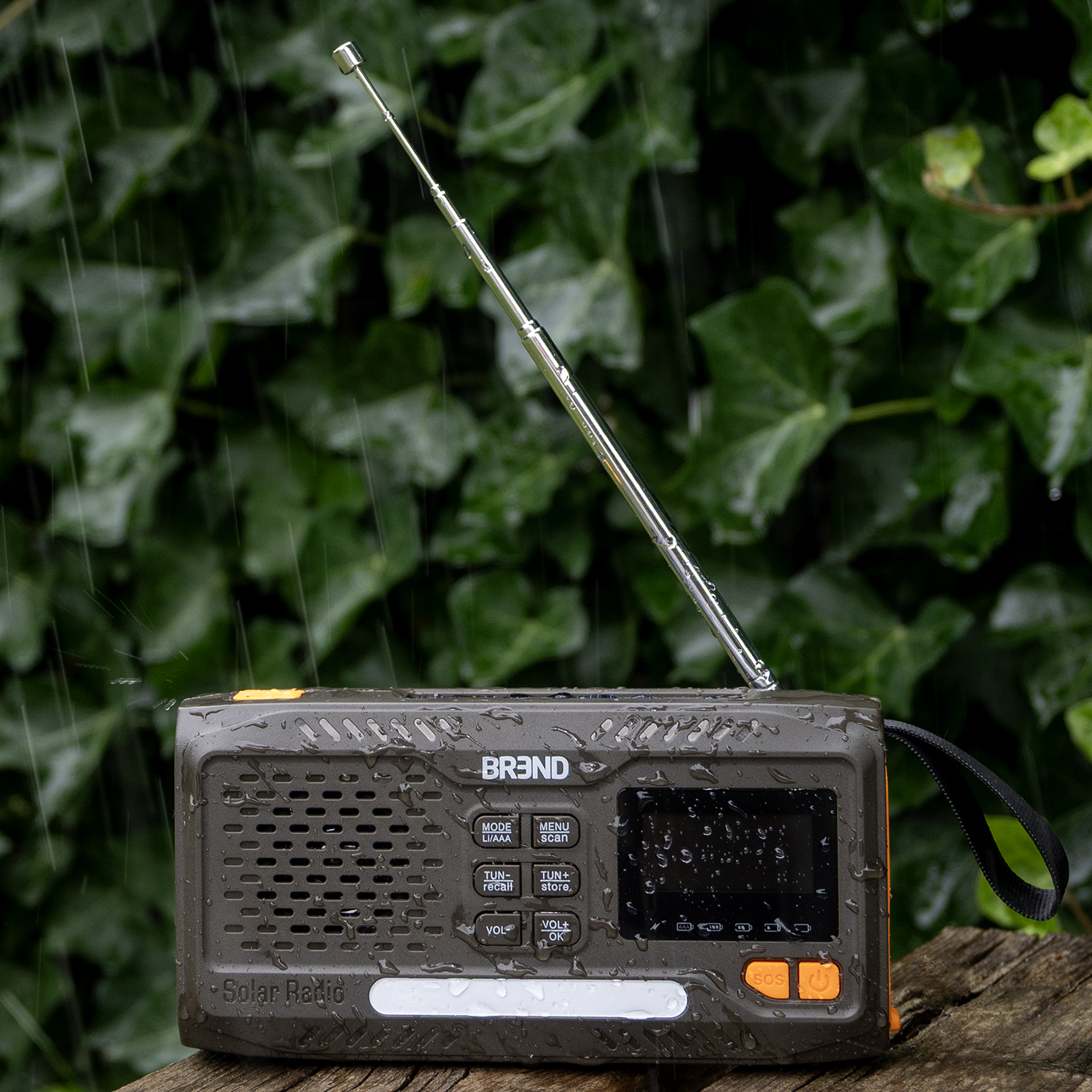 A solar radio sits on wood, water droplets on it. Green ivy is in the background, rain falling. Radio has antenna and orange accents.