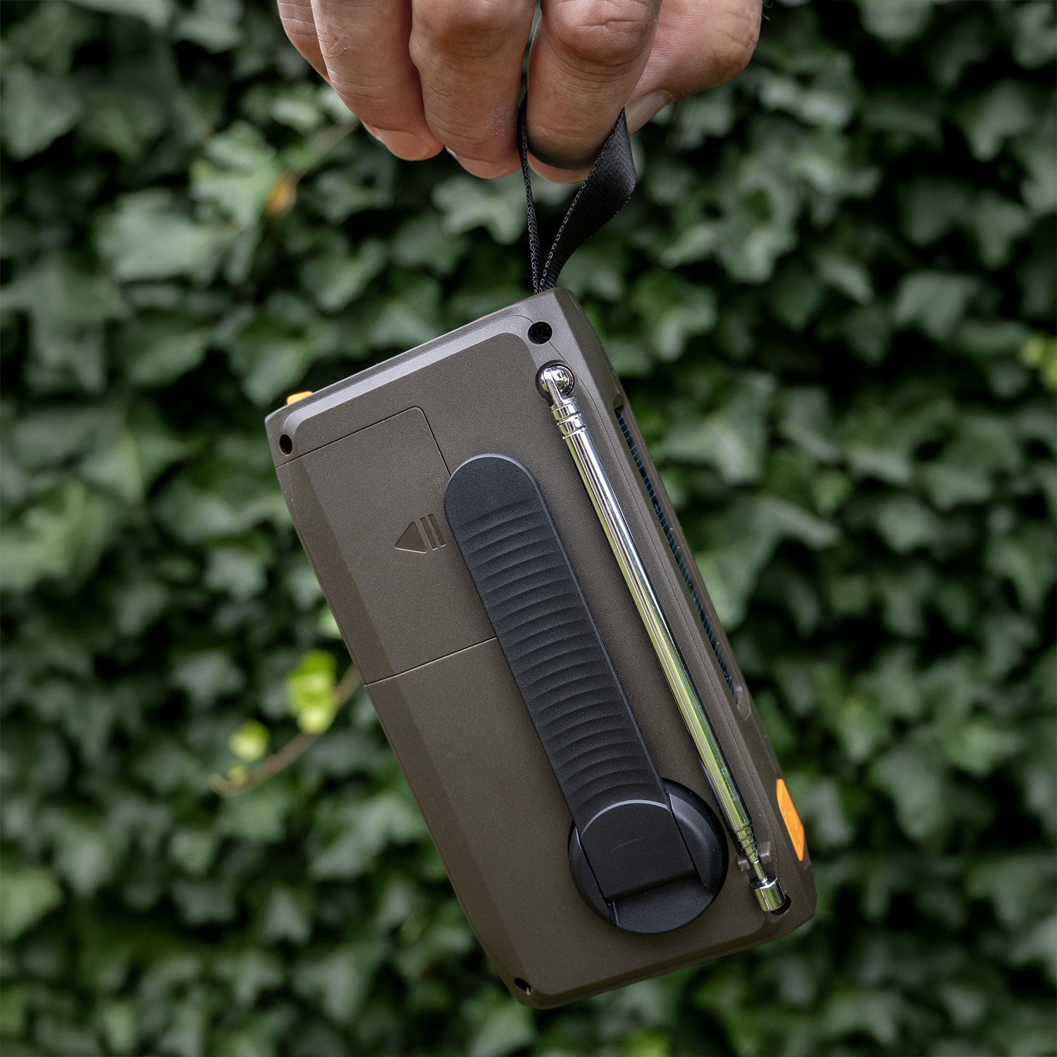 Hand holding a dark green radio with a black strap, antenna extended, against a blurred green foliage background.