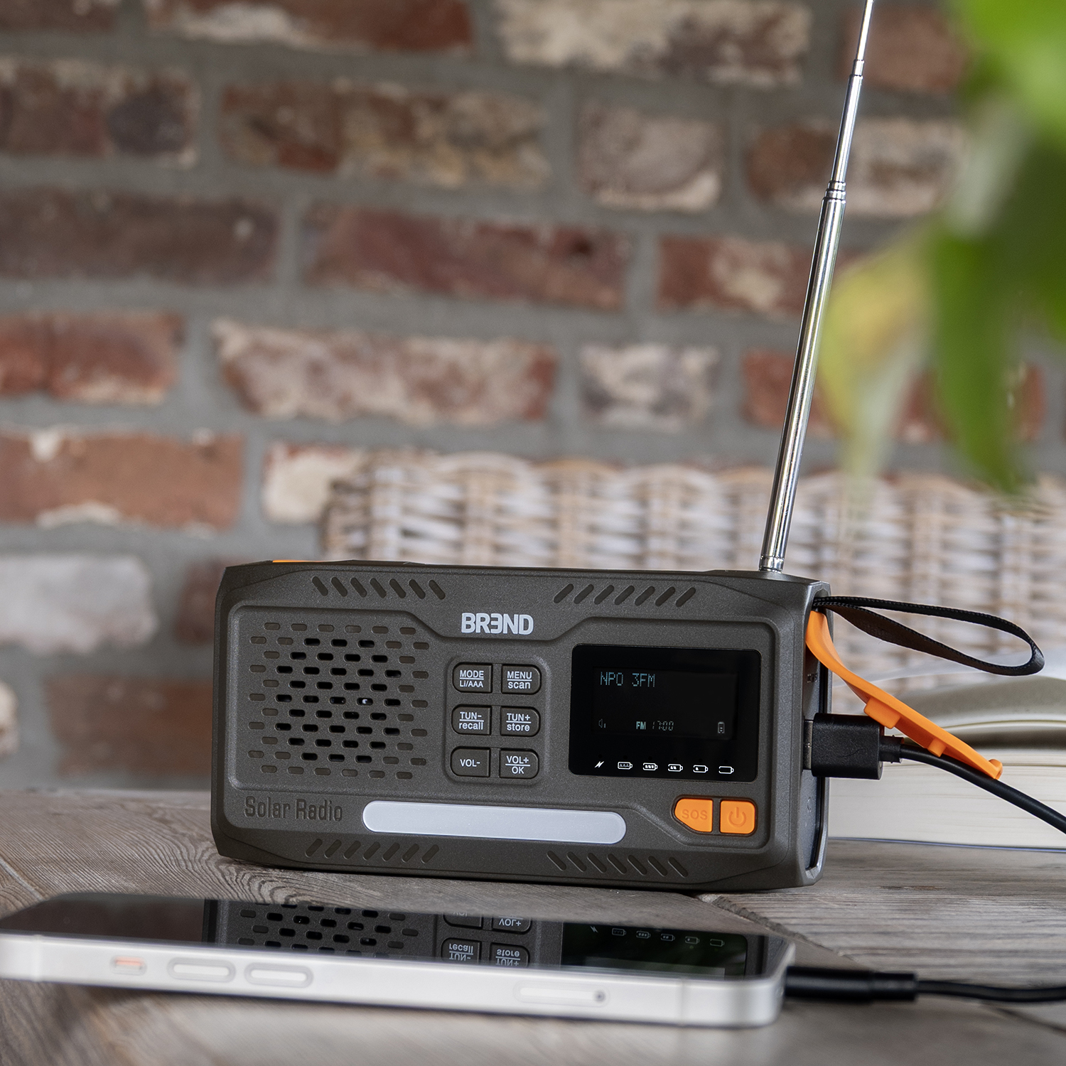 A solar radio with an antenna and a phone charging on a wooden table. A brick wall is in the background.