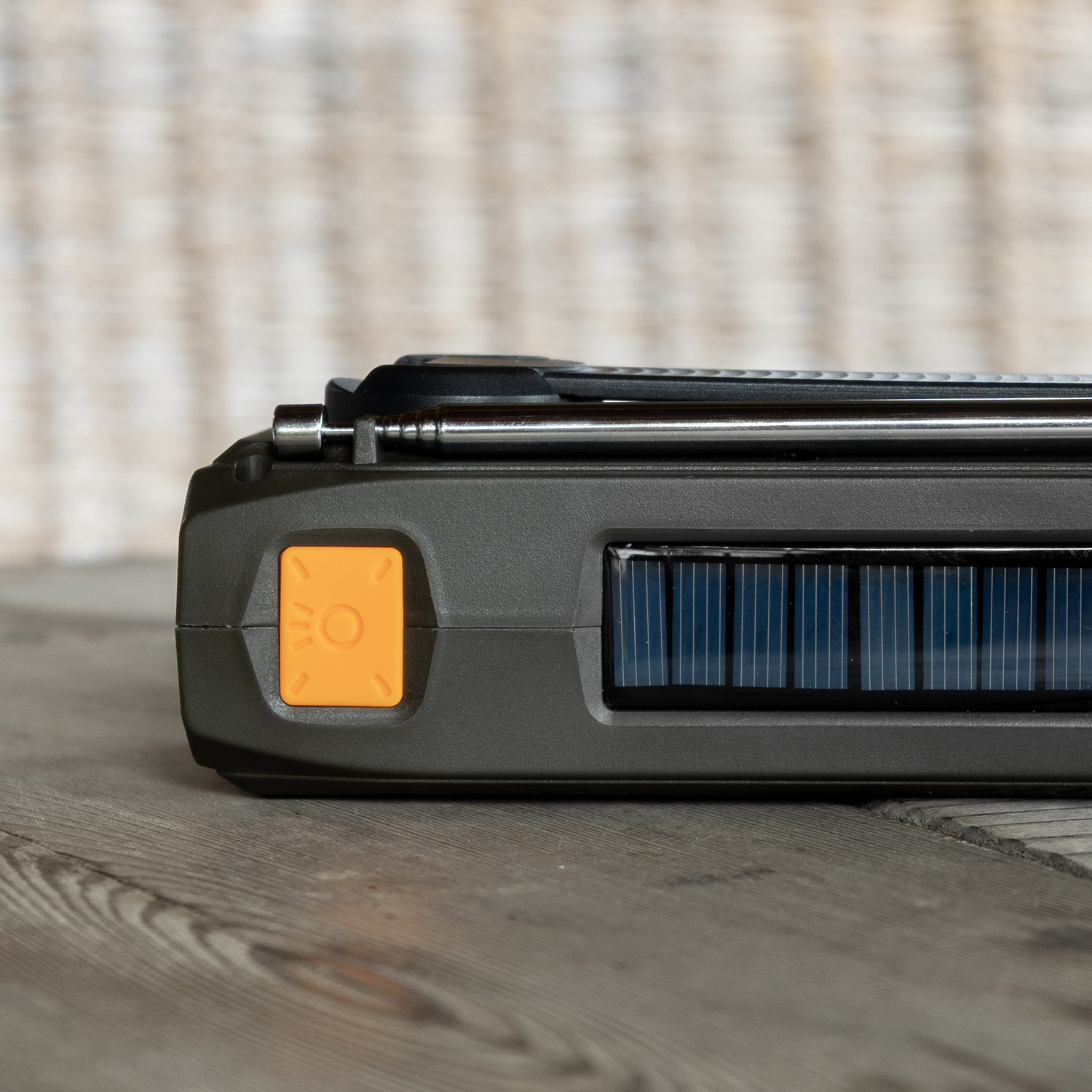 A close-up of a dark gray solar-powered radio with an orange button, a solar panel, and a silver antenna, on wooden surface.