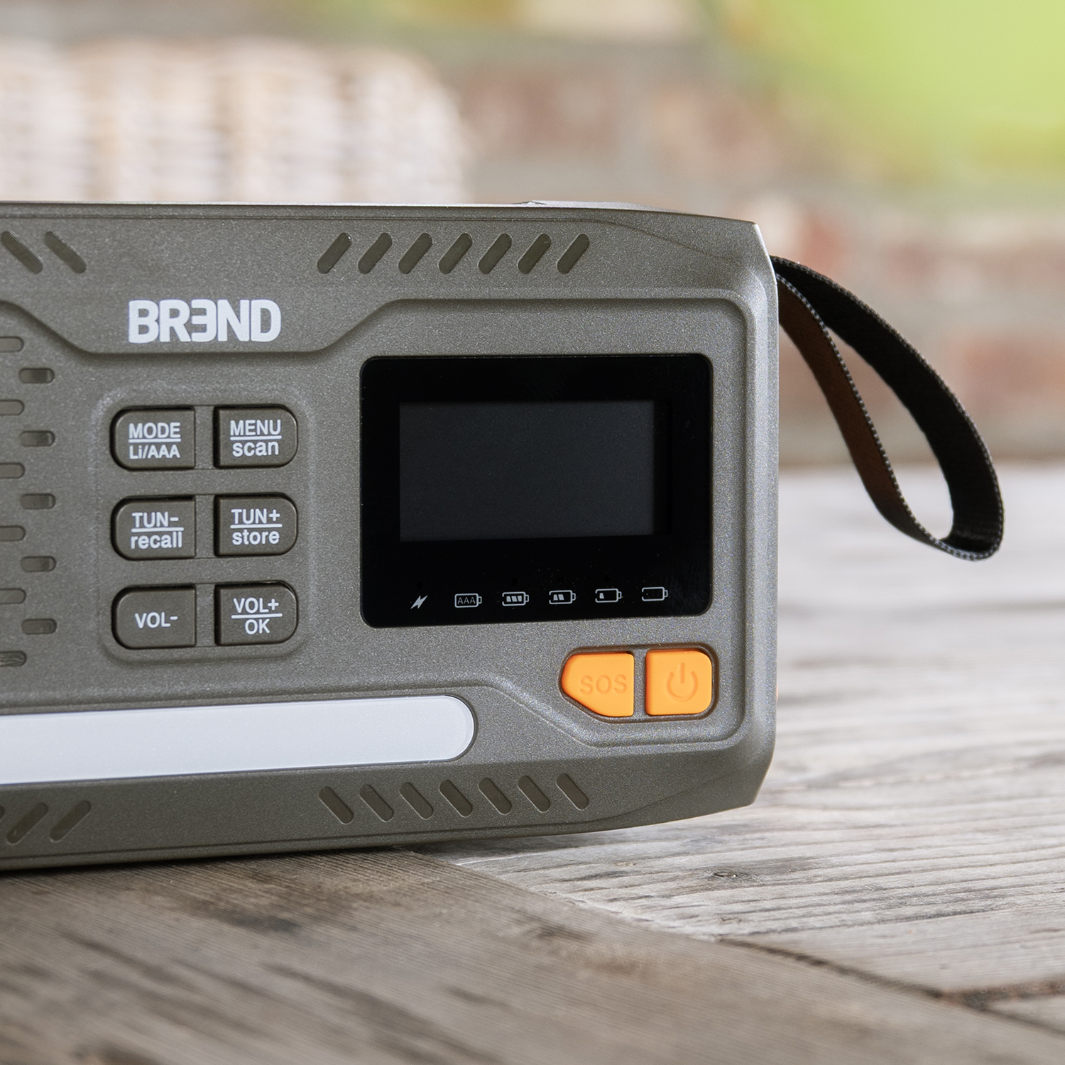 A close-up of a gray radio with buttons, an LCD screen, and a black strap. The radio has the brand name 'BR3ND' on it.