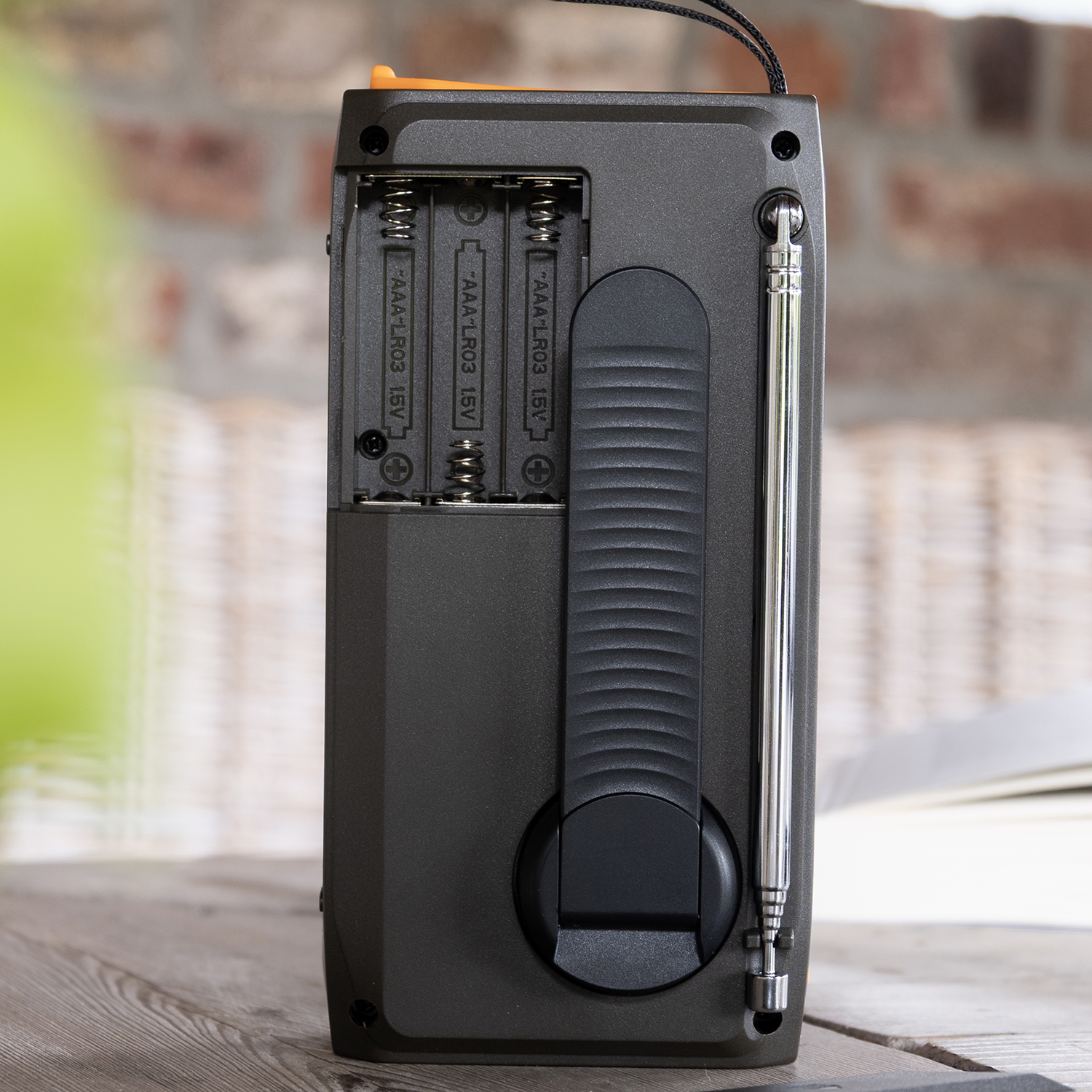 Close-up of a grey radio with antenna extended. Three AAA batteries are visible. The background has a blurred brick wall and wooden surface.