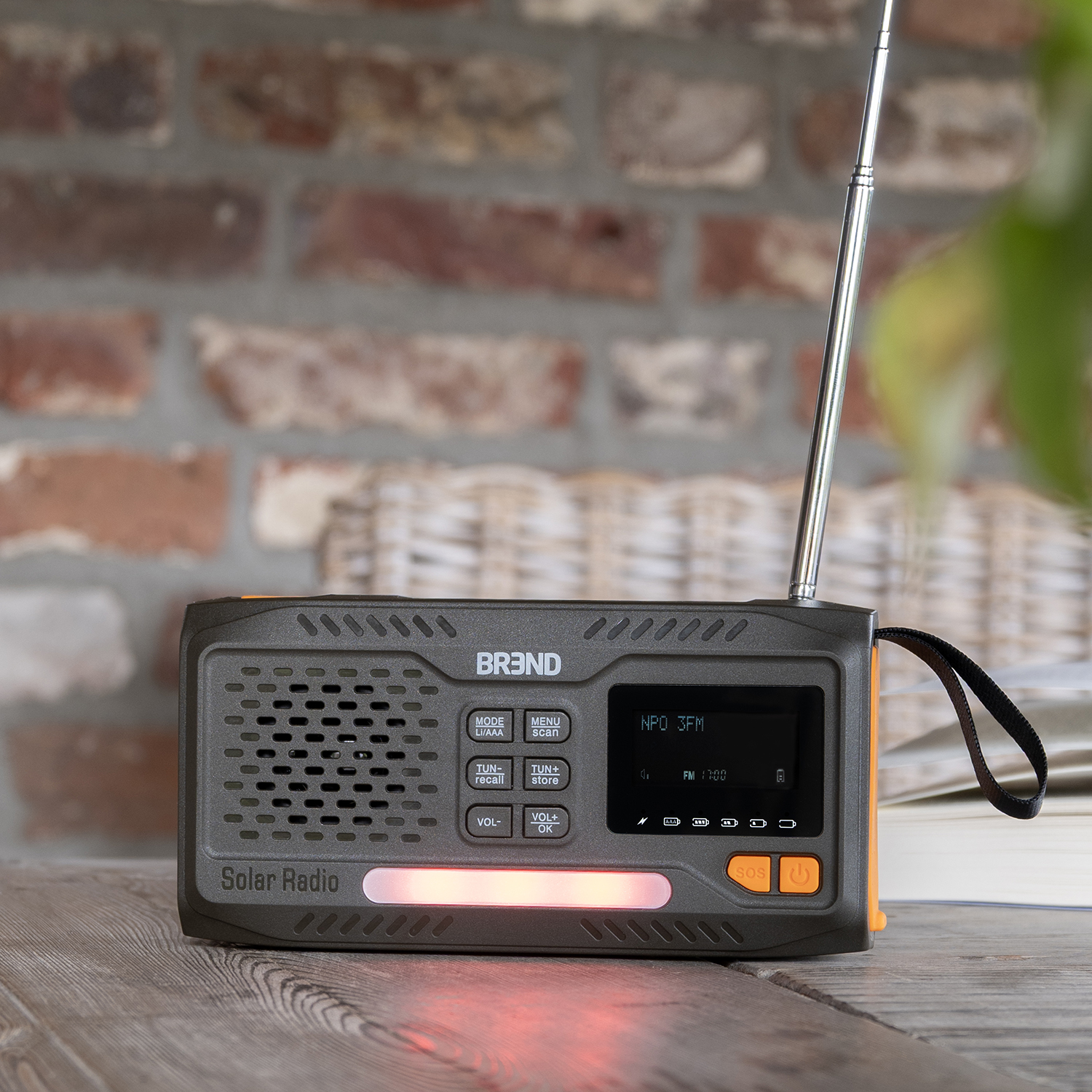 A solar radio sits on a wooden table. It is dark gray with orange accents. A brick wall and greenery are in the background.