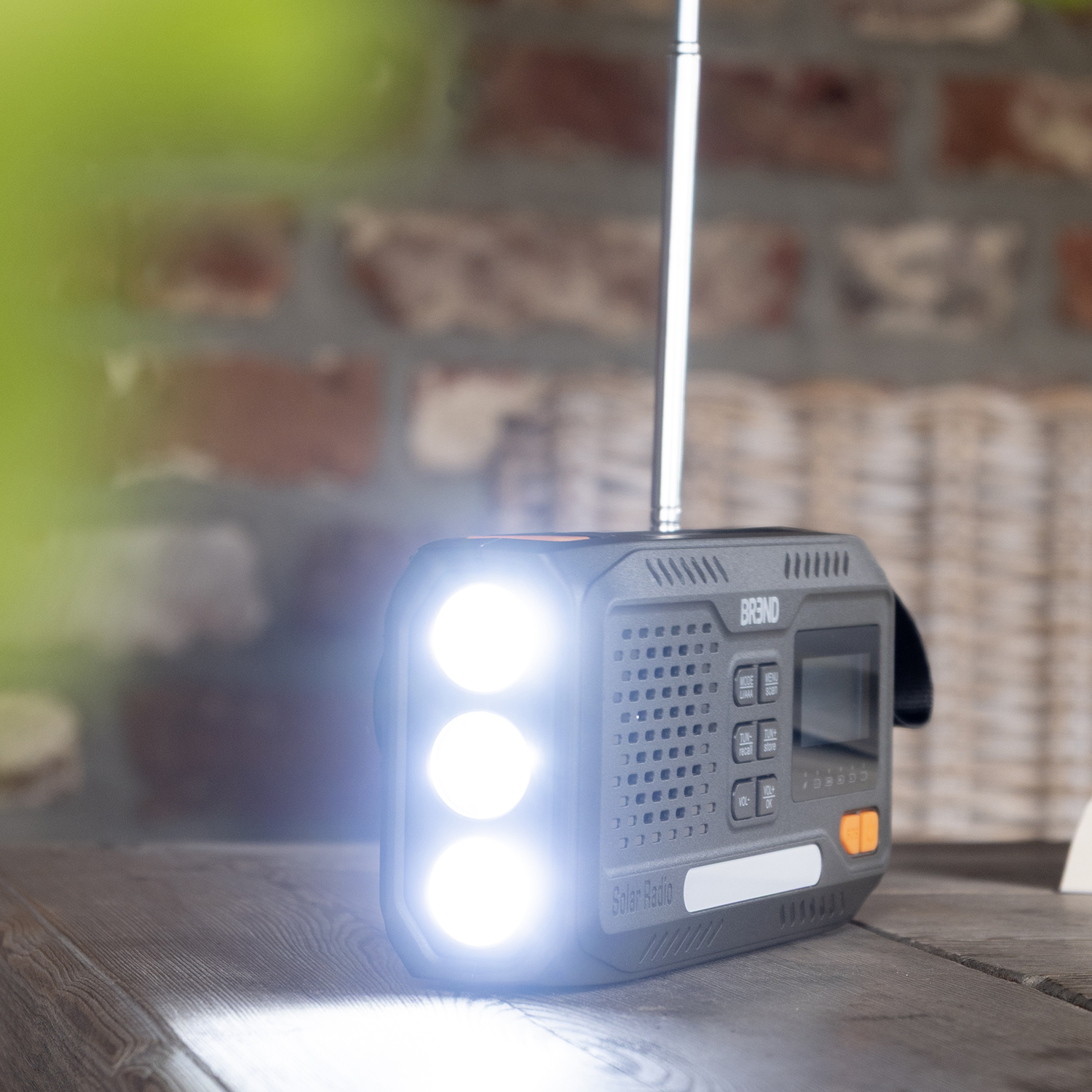 A solar radio with a bright flashlight is on a wooden table. In the background, there is a brick wall and wicker basket.