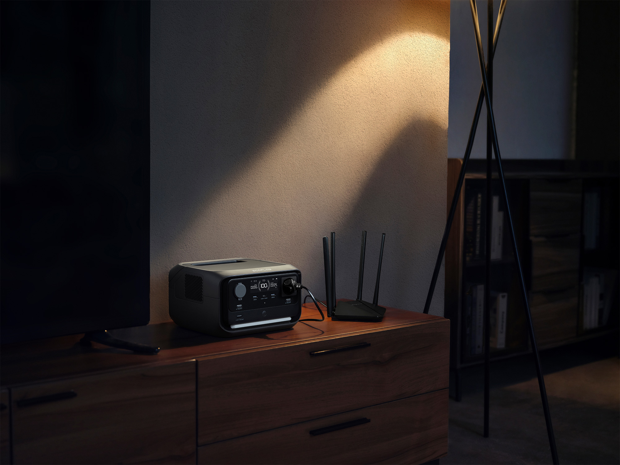 Eine schwarze Powerstation und ein Router stehen auf einem Holzschrank. Links ist ein Fernseher. Rechts steht eine Lampe.