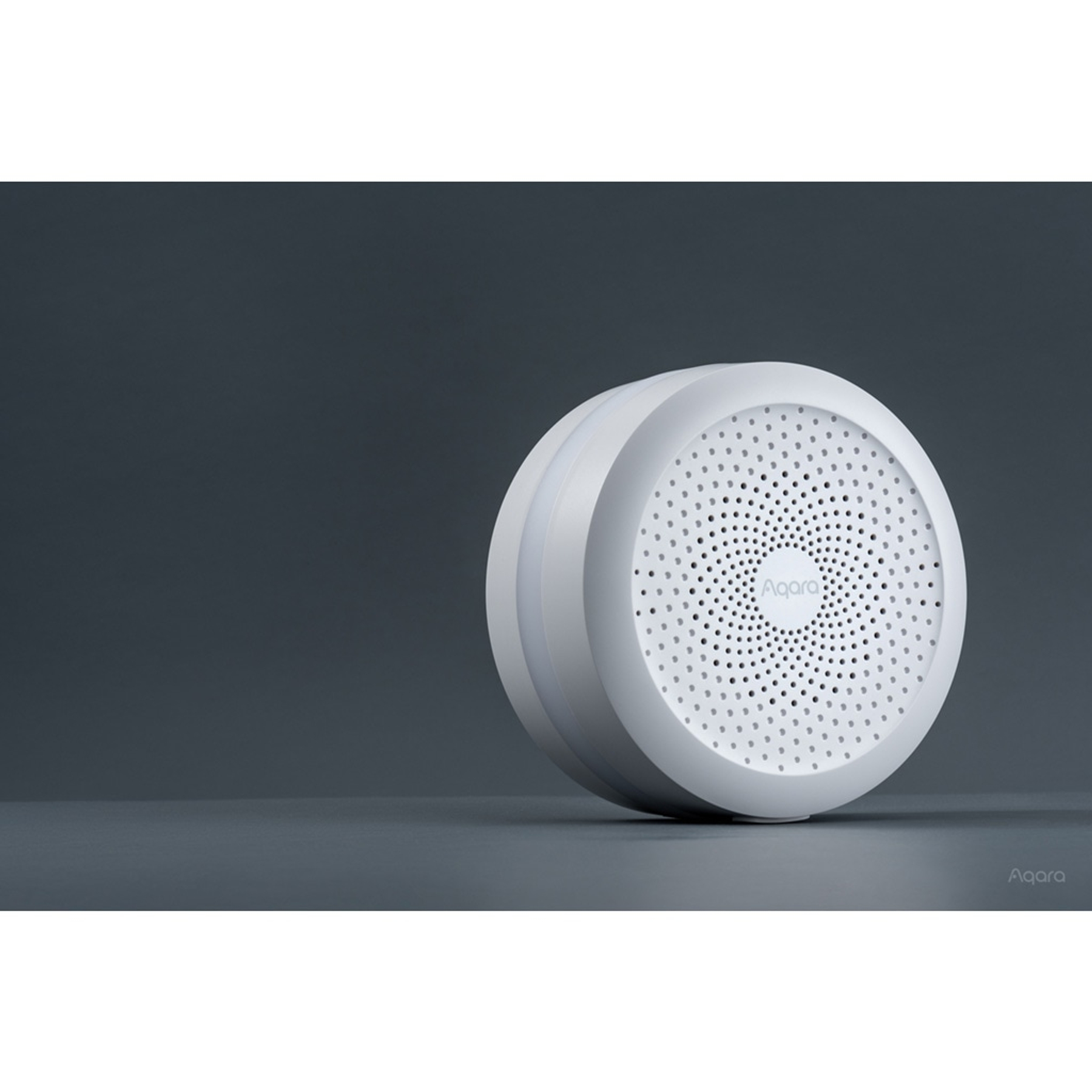 Een witte, cirkelvormige Aqara smart home-apparaat met veel kleine gaatjes aan de voorkant. Het staat op een grijs oppervlak.