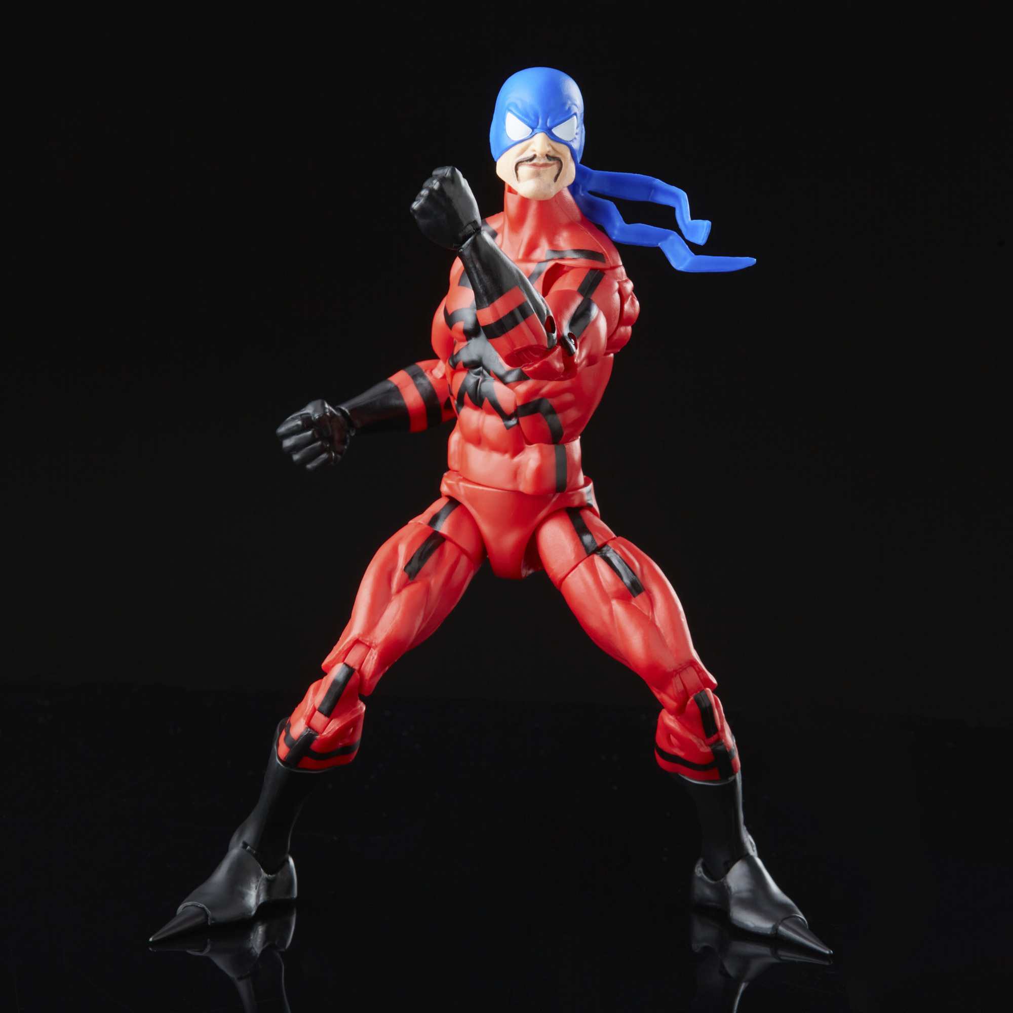 Una figura de superhéroe rojo con rayas negras y tocado azul posa contra un fondo negro, puños cerrados.