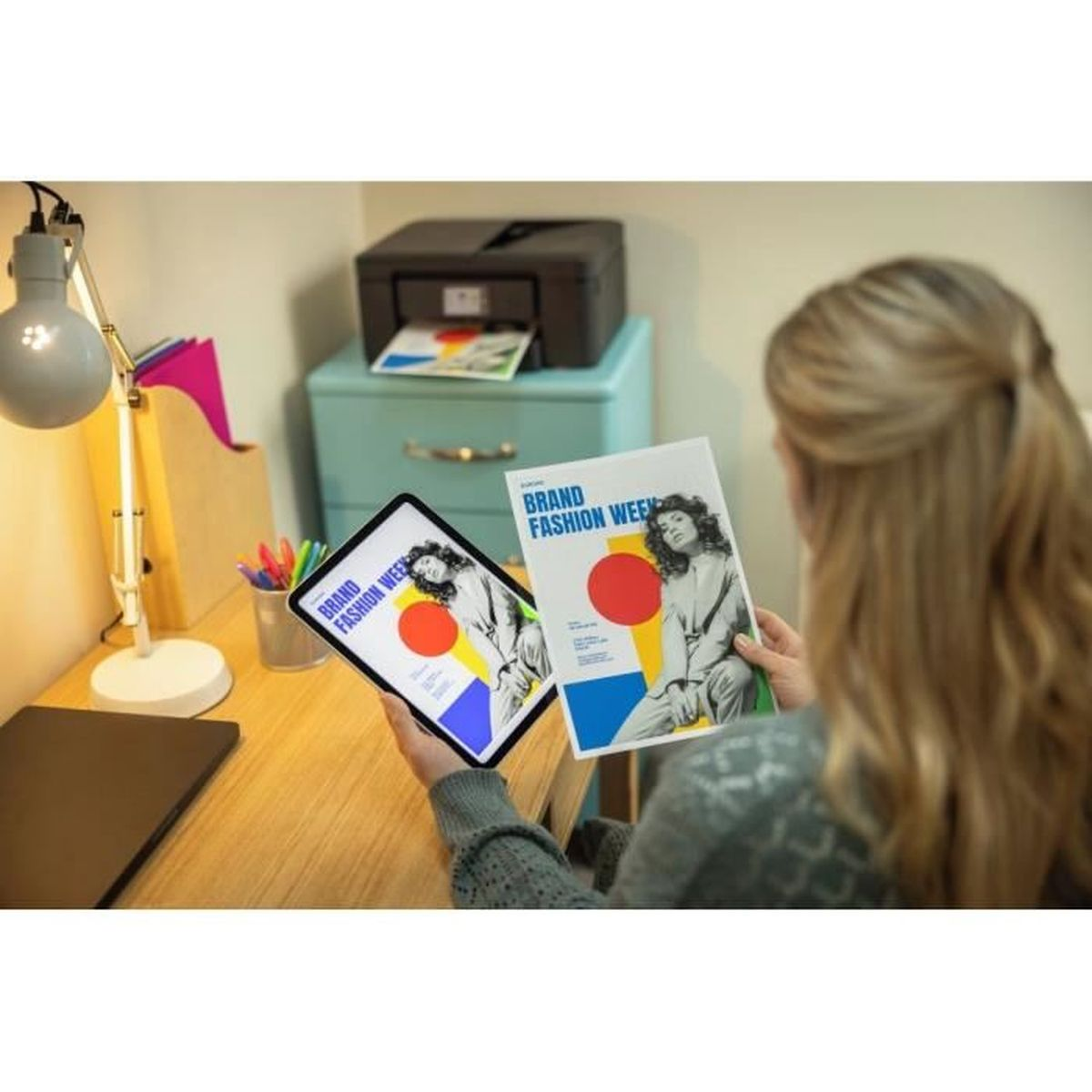 Kobieta trzyma tablet i ulotkę, oba z projektem 'Brand Fashion Week'. Widać również drukarkę i lampę biurkową.