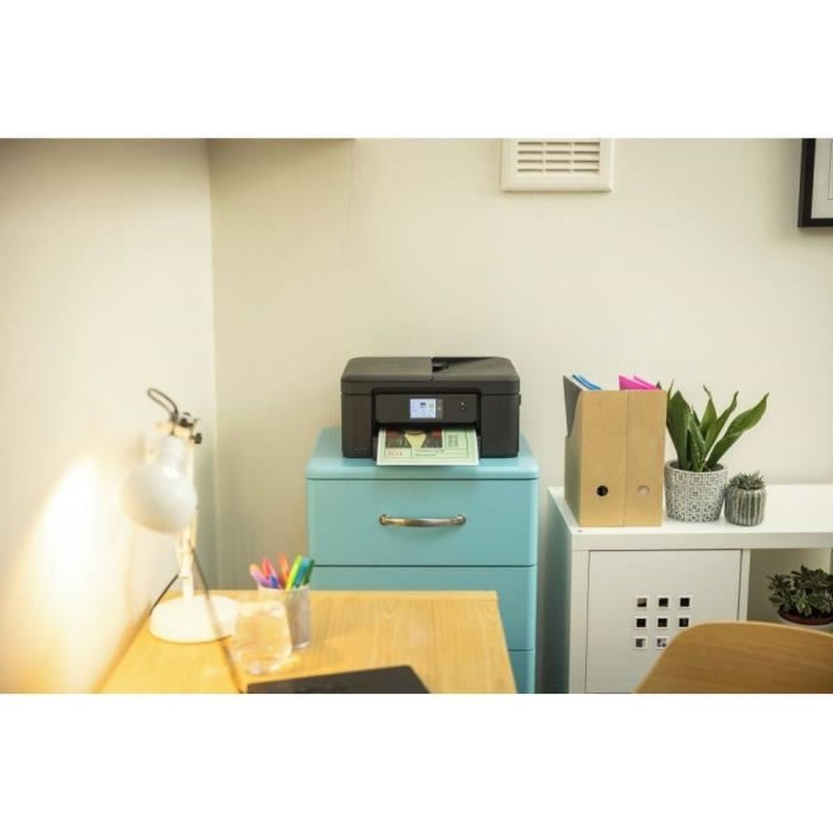 Czarna drukarka stoi na jasnoniebieskiej szafce. Widoczny wydrukowany dokument. Biurko, lampa i roślina również w pokoju.