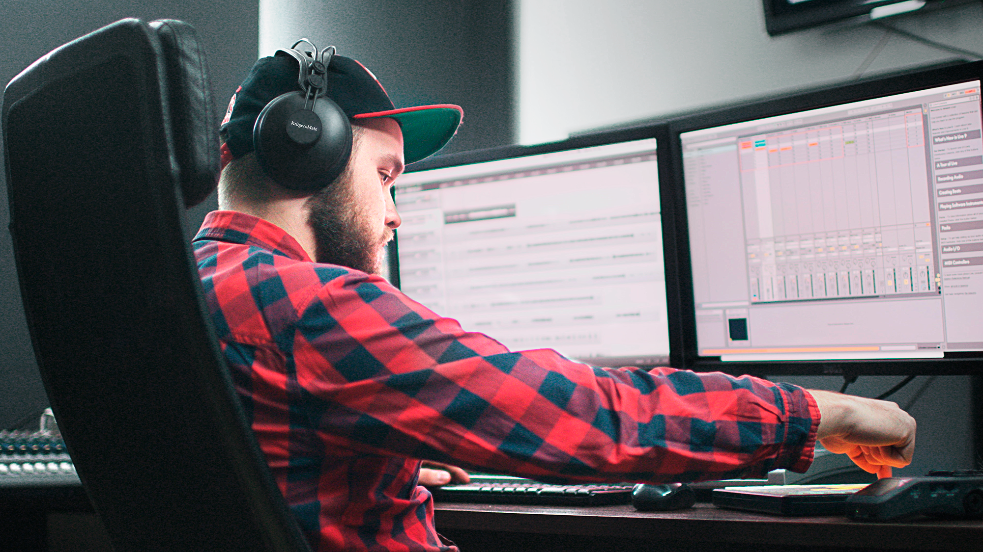 Mężczyzna w czerwonej koszuli w kratę, w słuchawkach, pracuje przy komputerze. Ma przed sobą dwa monitory.