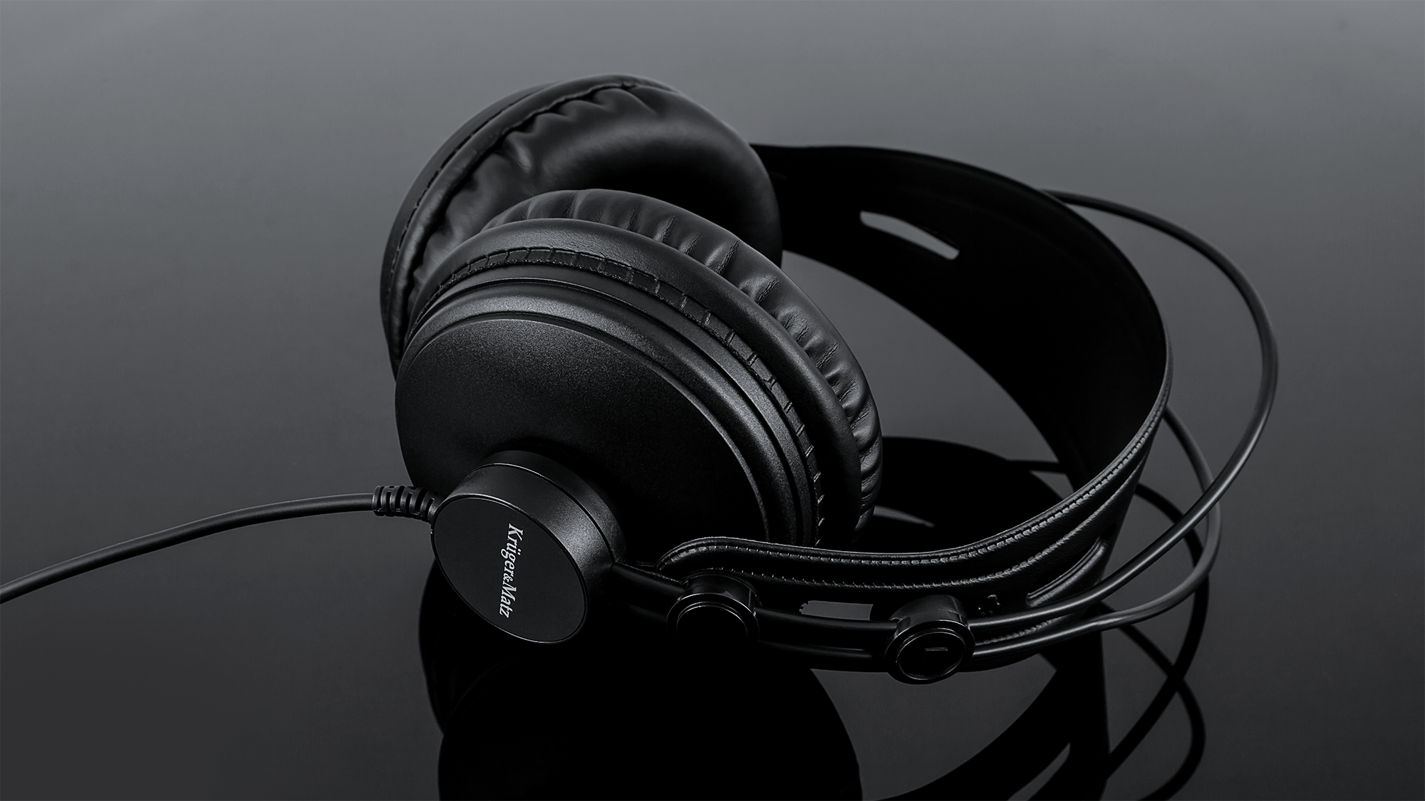Czarne słuchawki nauszniki na odbijającej powierzchni. Słuchawki oznaczone jako 'Krüger&Matz'.