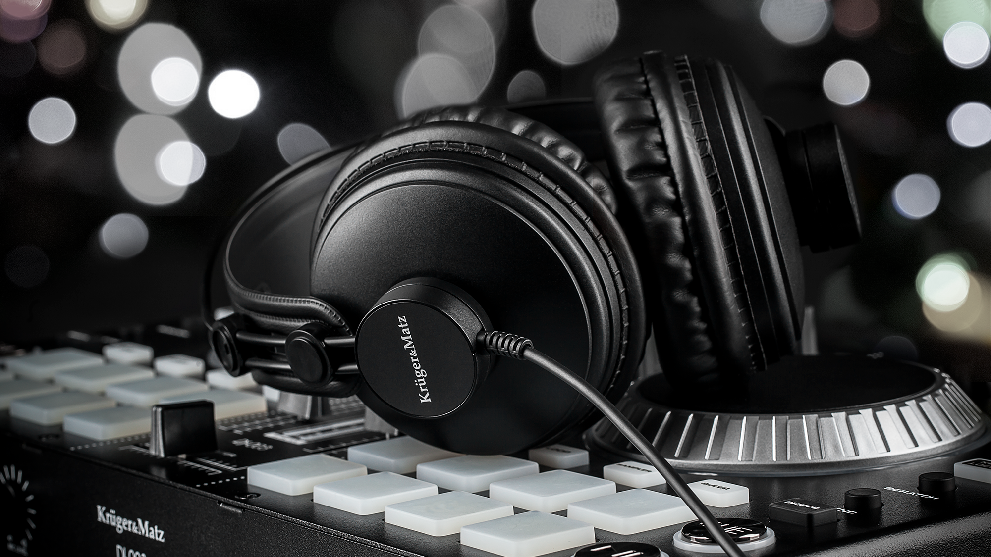 Czarne słuchawki spoczywają na kontrolerze DJ. Tło ma rozmyte światła. Kontroler ma białe pady i przyciski.