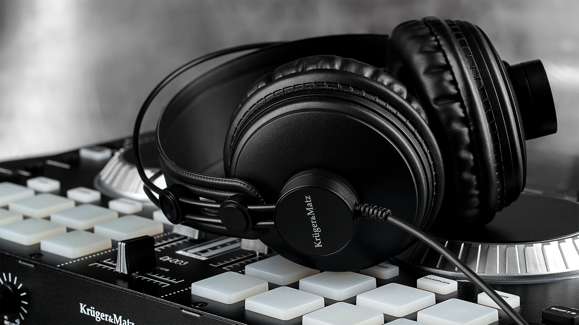 Czarne słuchawki spoczywają na mikserze DJ. Słuchawki mają styl nauszny. Mikser ma białe kwadratowe pady. Tło jest rozmyte.