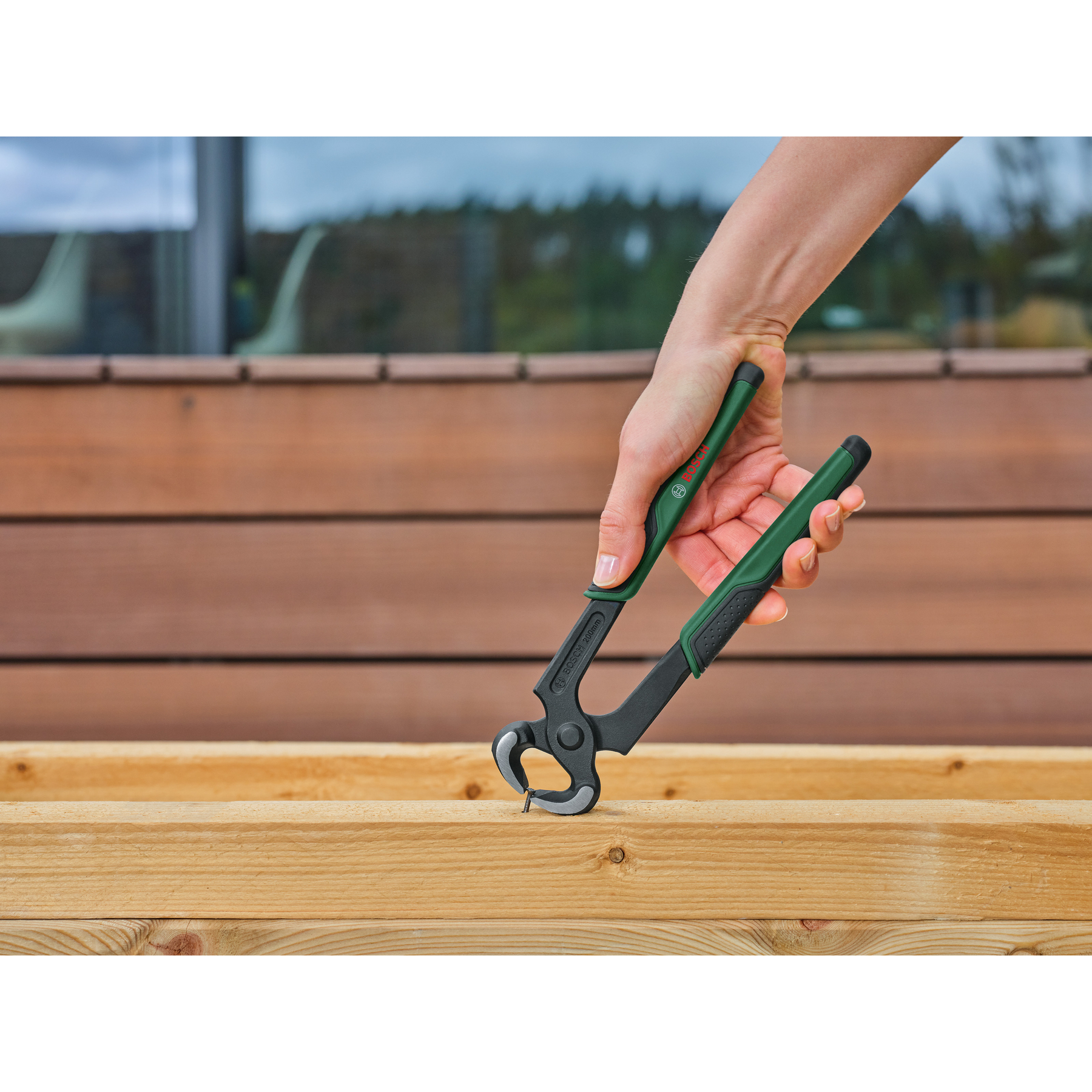 Hand hält eine grün-schwarze Zange über Holz mit einem Nagel. Hintergrund zeigt Holzbretter und ein Fenster.