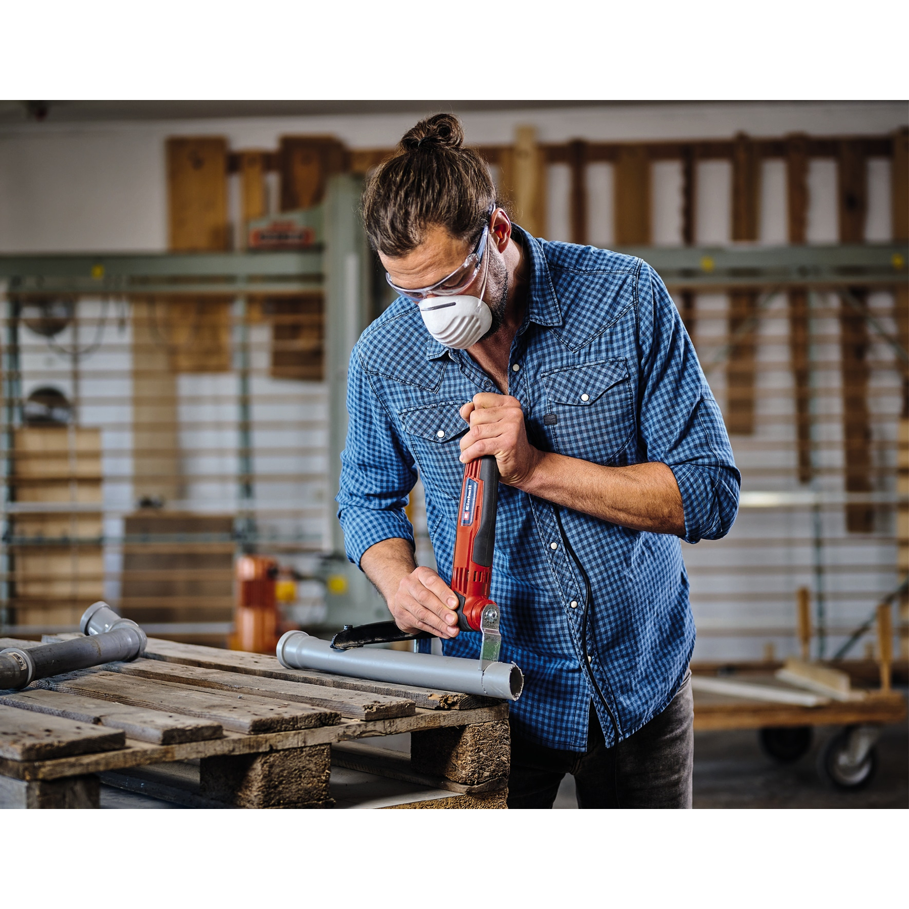 Mężczyzna w okularach i masce tnie szarą rurę czerwono-czarnym narzędziem. Rury leżą na drewnianej palecie.
