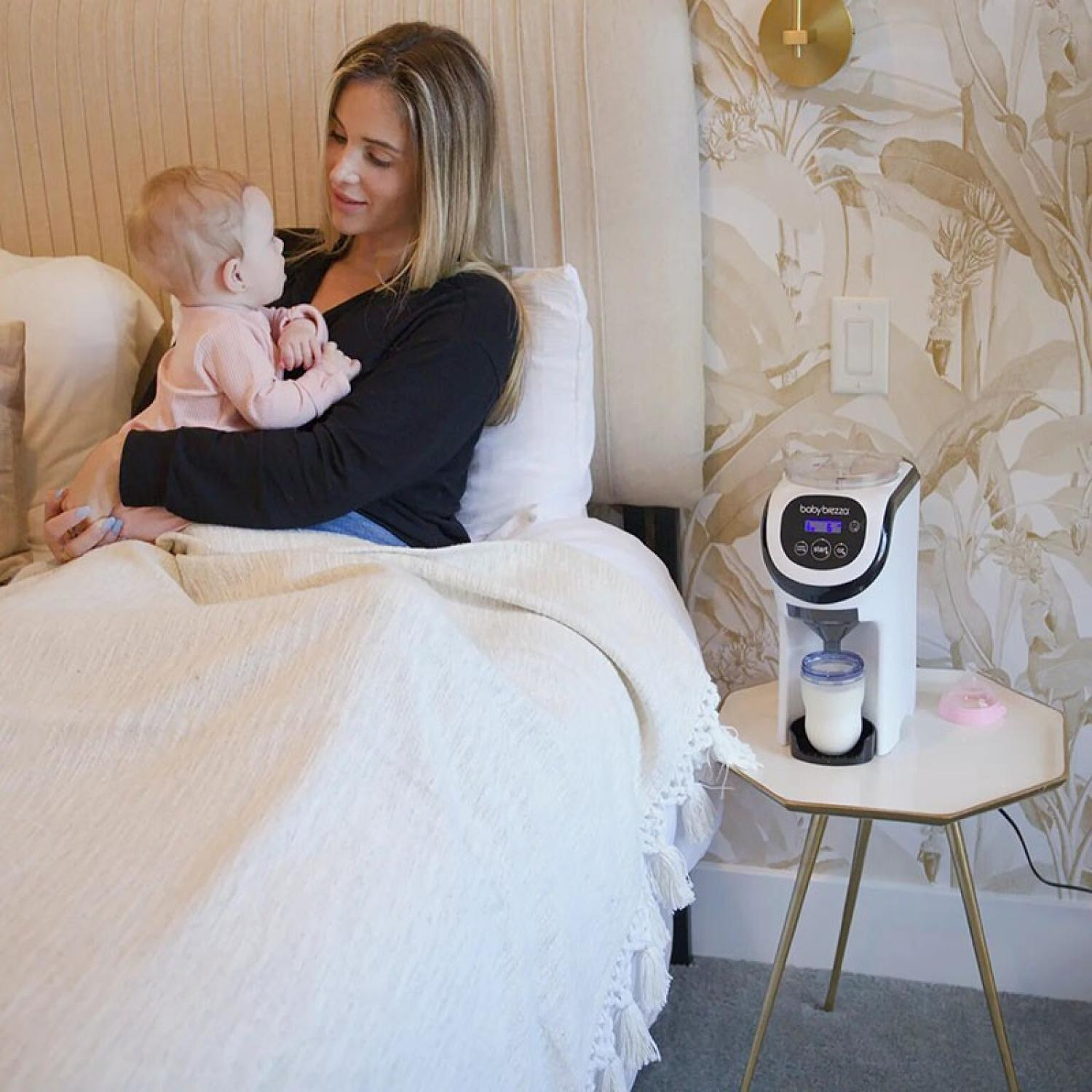Eine Frau hält ein Baby auf einem Bett. Ein Formelzubereiter steht auf einem Beistelltisch. Das Zimmer hat Blumentapeten und eine Wanduhr.
