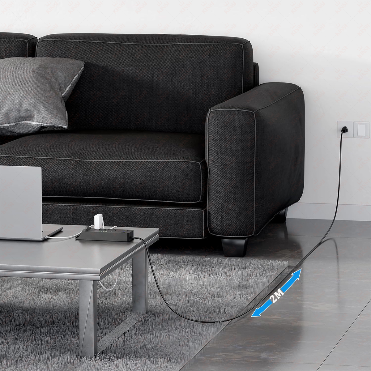 Ein schwarzes Sofa mit einem grauen Kissen, ein silberner Tisch mit einem Laptop und eine Steckdosenleiste. Ein Kabel führt zu einer Steckdose.