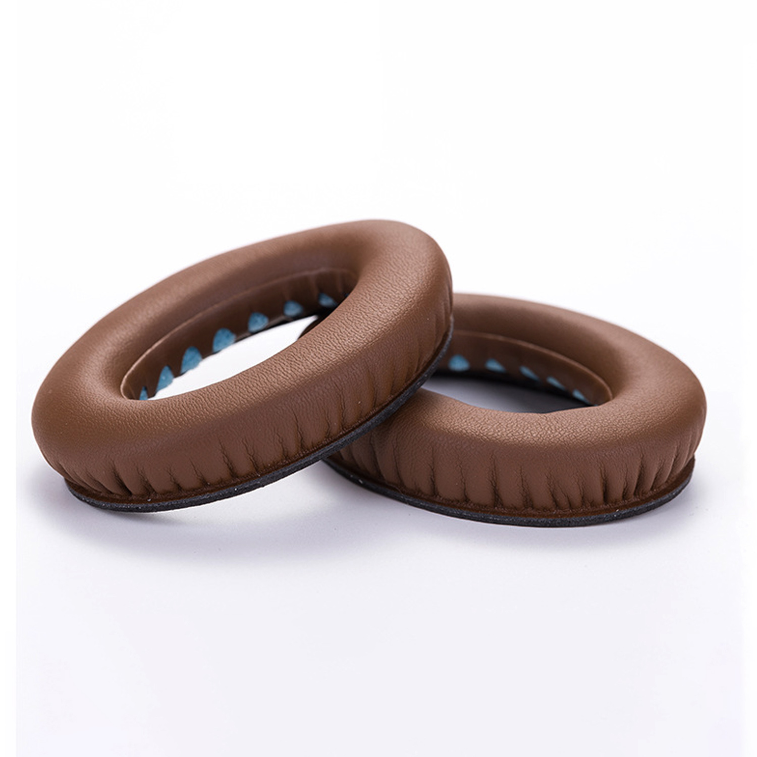 Dwie brązowe, owalne poduszki na uszy do słuchawek, leżące na białej powierzchni. Jedna lekko nachodzi na drugą.