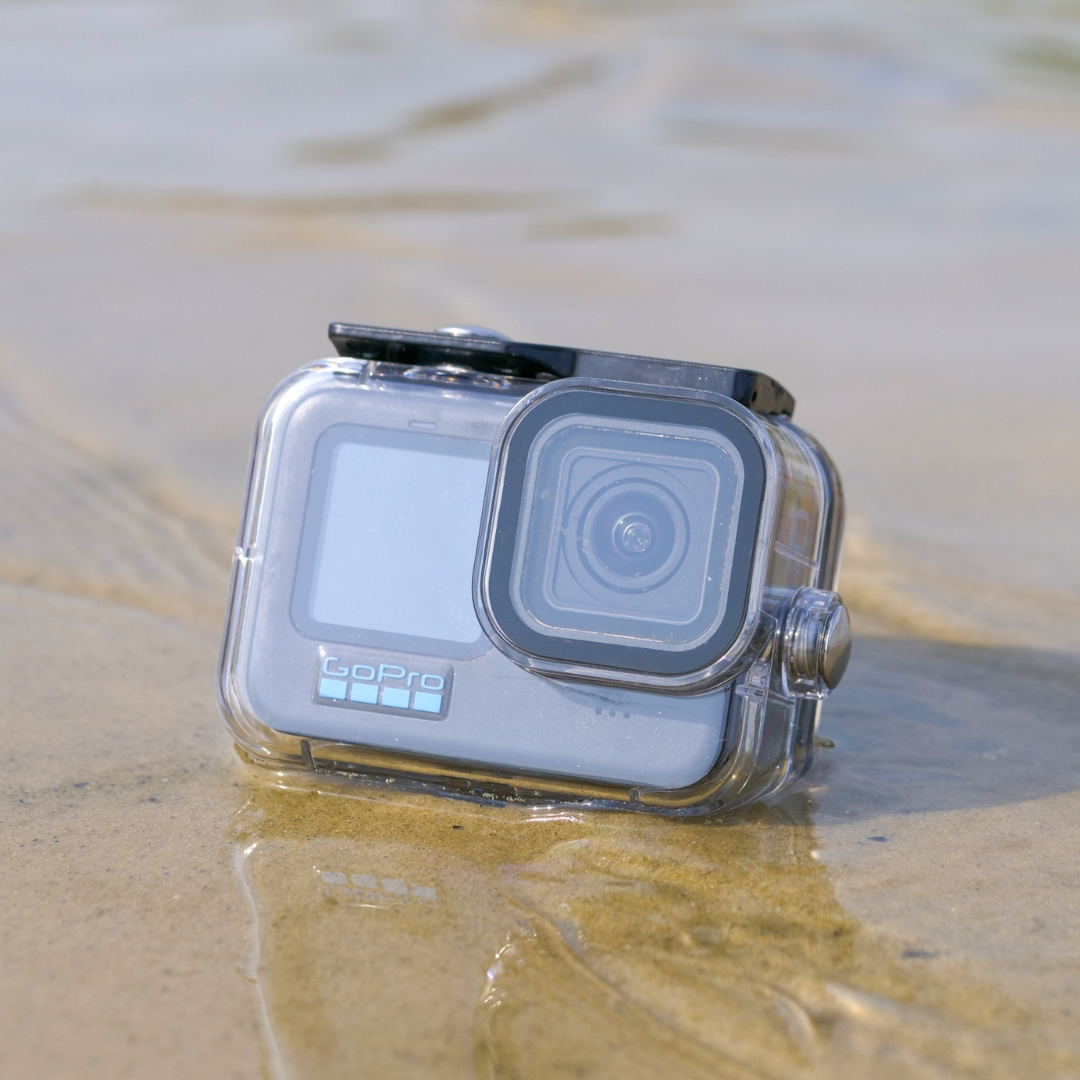 Een GoPro camera in een doorzichtige waterdichte behuizing ligt op nat zand. De camera is grijs en zwart met een scherm en lens zichtbaar.