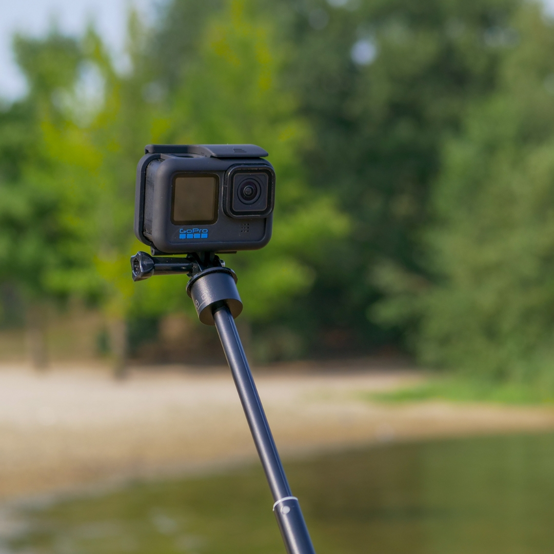 Eine schwarze GoPro-Kamera ist auf einem Selfie-Stick montiert. Der Hintergrund umfasst grüne Bäume, ein sandiger Ufer und Wasser.