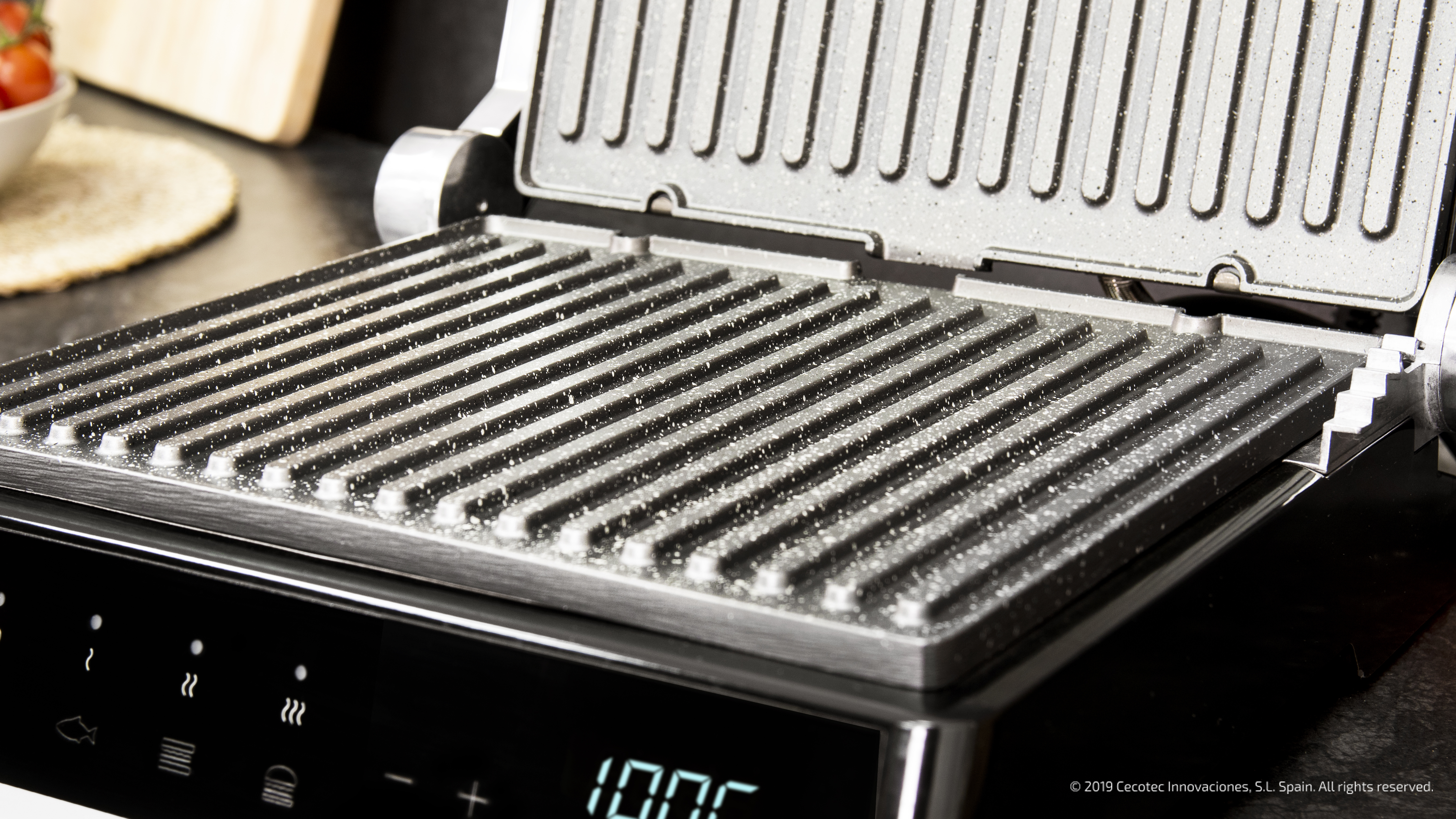 Otwarty grill z czarną powierzchnią, pokazujący równoległe linie. Wyświetlacz cyfrowy pokazuje temperaturę. Pomidory widoczne w tle.