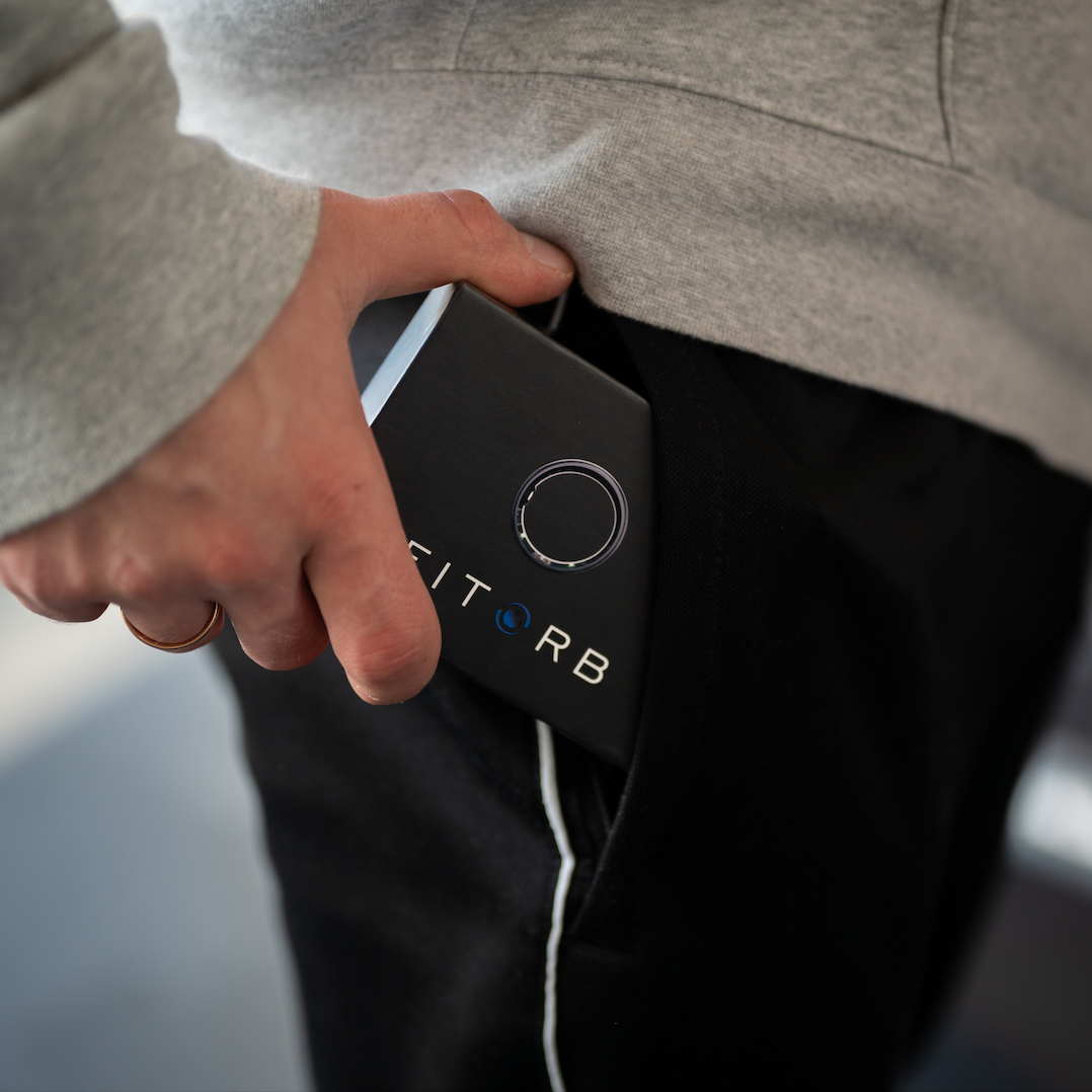 Eine Person steckt ein schwarzes Gerät mit der Aufschrift FITORB und einem Kreisdesign in eine Hosentasche. Ein grauer Kapuzenpullover ist ebenfalls zu sehen.