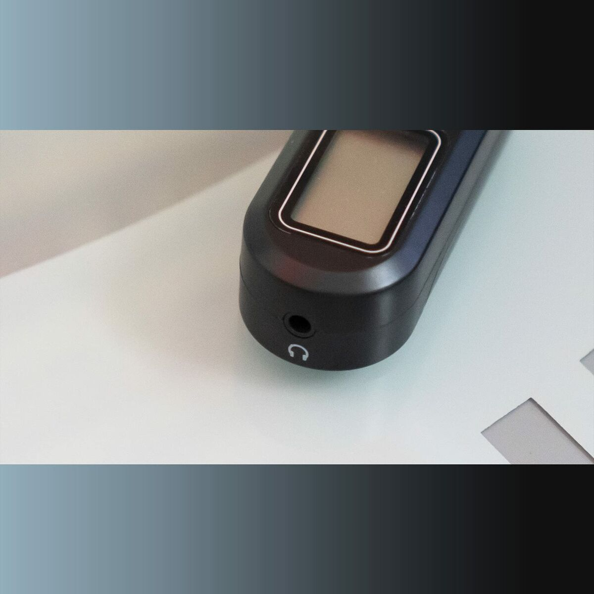 Czarne urządzenie elektroniczne z ekranem leży na białej powierzchni. Ma gniazdo słuchawkowe. Tło to gradient.