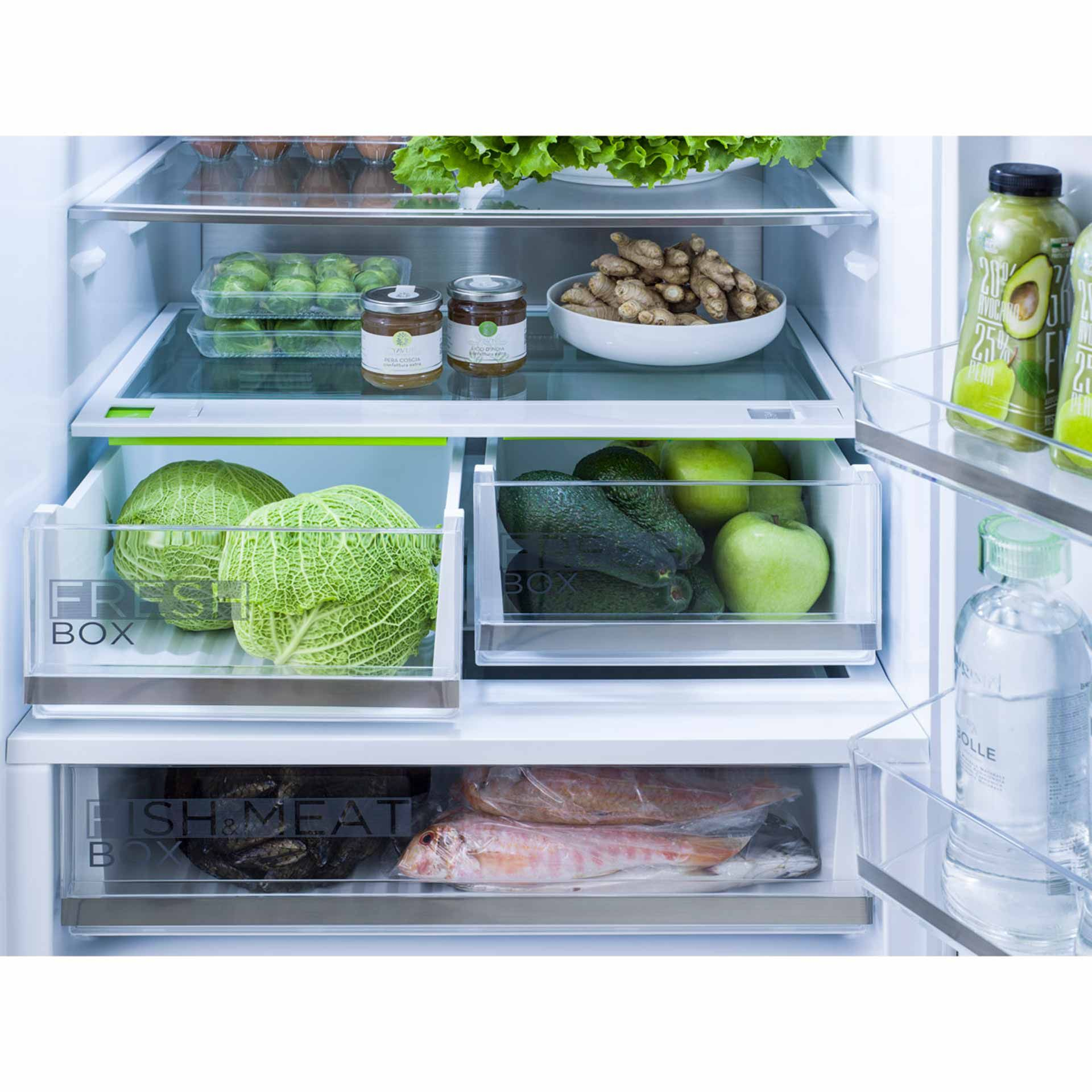 Interno del frigorifero pieno di prodotti freschi: cavoli, avocado, mele, zenzero, barattoli, bevande in bottiglia e pesce.