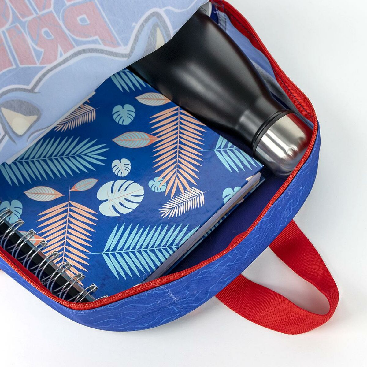 Ein blauer Rucksack mit rotem Griff, der ein Notizbuch, eine schwarze Wasserflasche und einen hellblauen Stoff mit Muster enthält.
