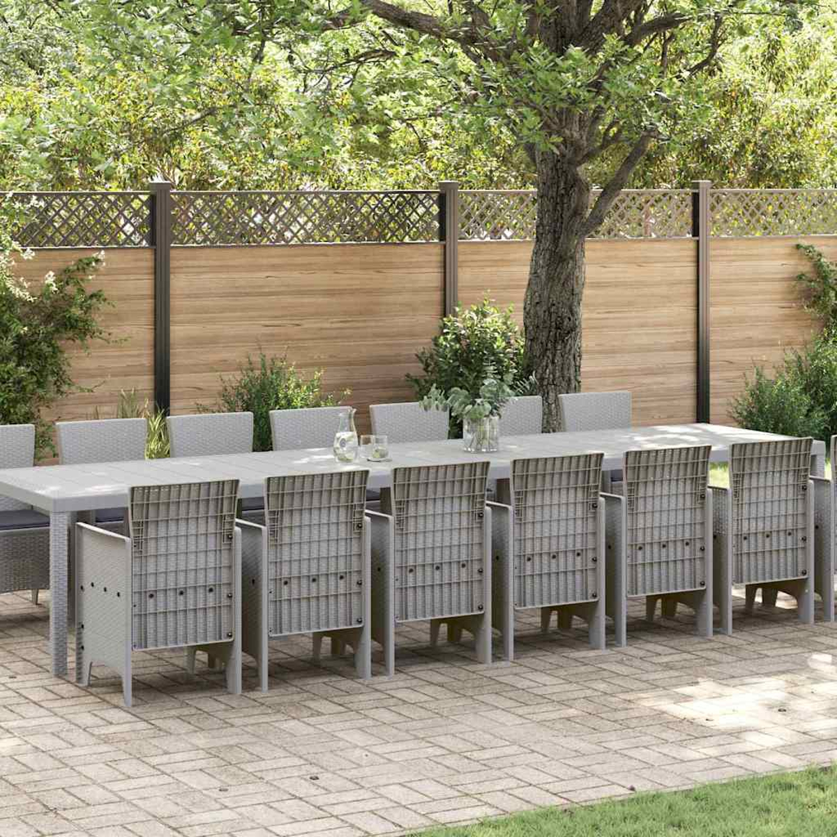 Ein Essbereich im Freien. Ein langer grauer Tisch mit 10 Stühlen steht auf einer Ziegelterrasse, im Hintergrund ein Holzzaun und Bäume.