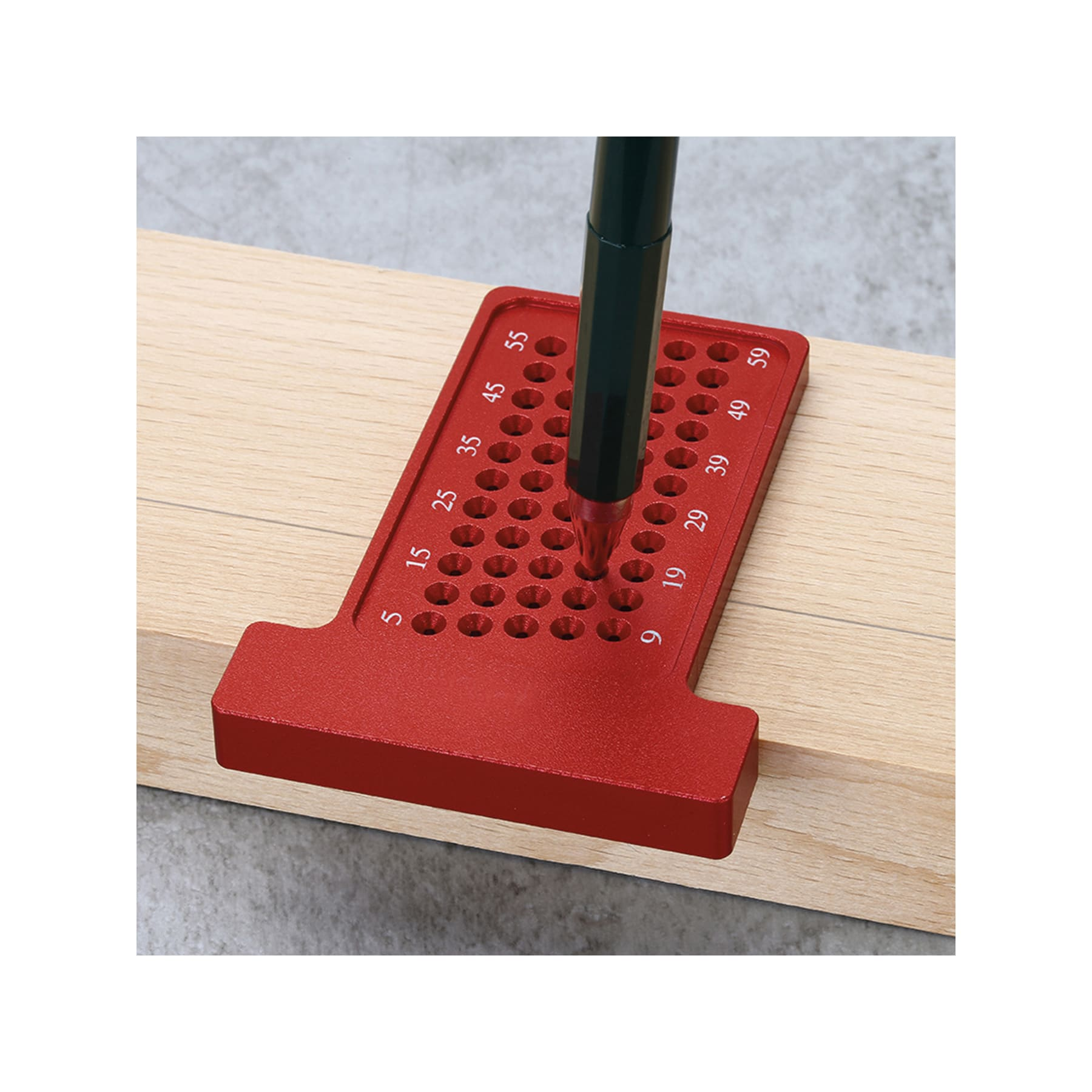 Ein rotes Metall-Markierungswerkzeug mit einem Stift liegt auf einem Holzbrett. Das Werkzeug hat Löcher zum Markieren. Der Hintergrund ist grau.