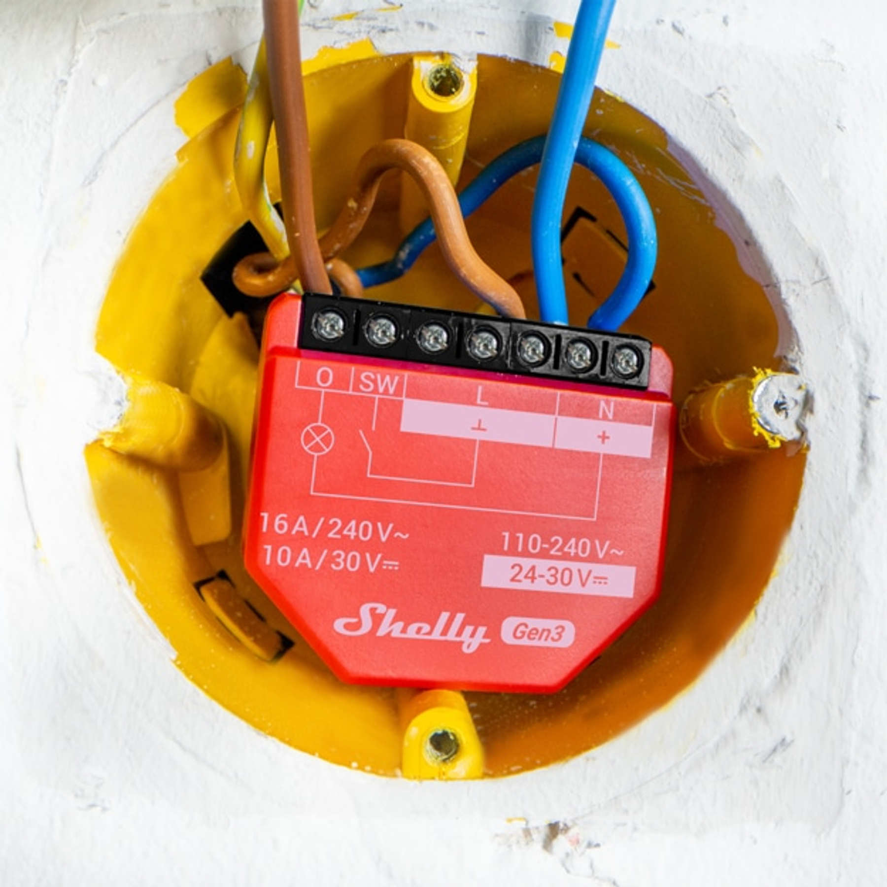 Czerwone urządzenie Shelly Gen3 z przewodami w żółtej wpuszczanej skrzynce elektrycznej. Przewody są brązowe, niebieskie i pomarańczowe na białej ścianie.