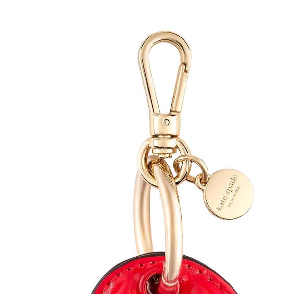 Ein goldfarbener Verschluss mit einem Kate Spade New York Anhänger. Der Verschluss ist an einem roten, glänzenden Objekt befestigt, wahrscheinlich eine Handtasche.