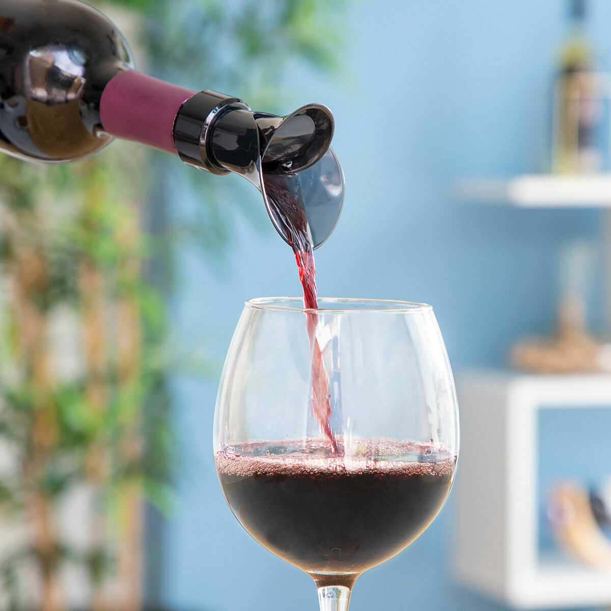 Rotwein wird aus einer Flasche mit einem Belüfter in ein Weinglas gegossen. Blaue Wand und grüne Pflanzen im Hintergrund.