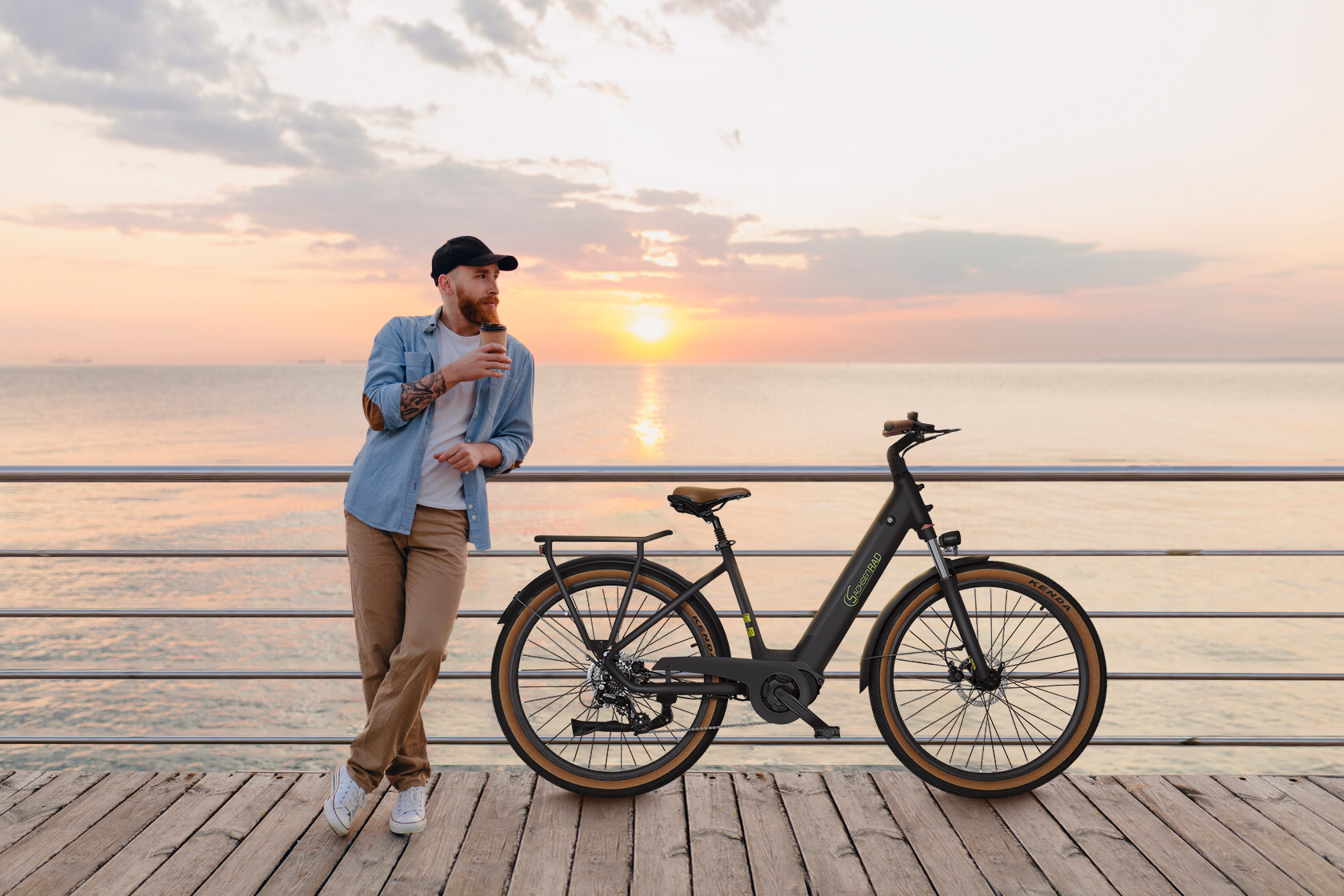 Ein Mann mit Bart lehnt an einem Geländer, hält Kaffee, neben einem Fahrrad auf einem Pier. Sonnenuntergang über dem Meer.