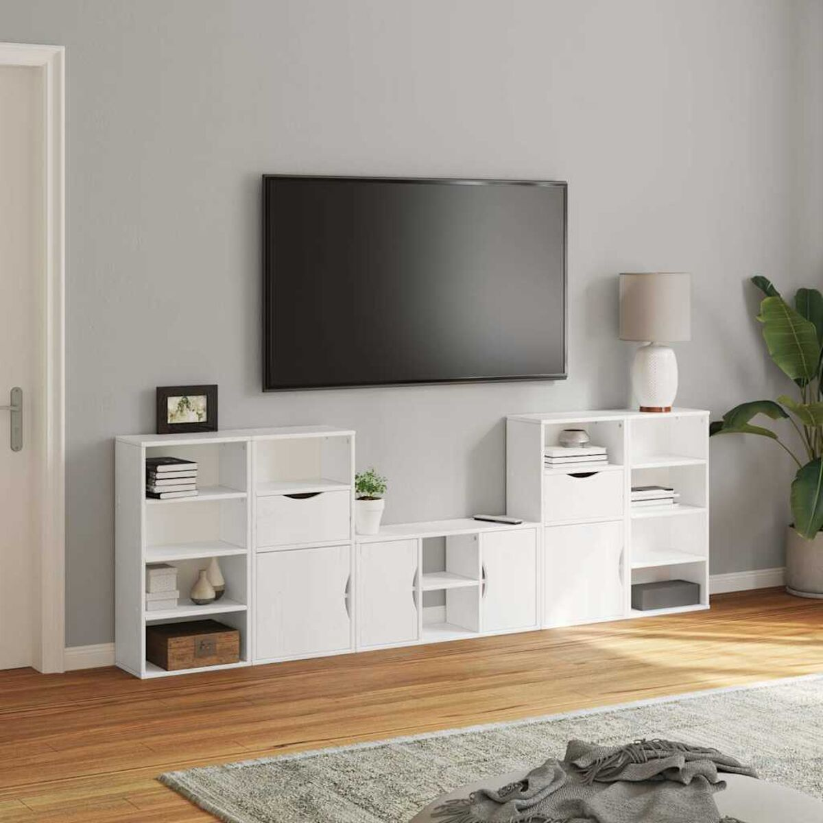 Ein Wohnzimmer mit großem Fernseher an einer grauen Wand. Weiße Schränke stehen darunter. Eine Pflanze, eine Lampe und eine Tür sind ebenfalls sichtbar.