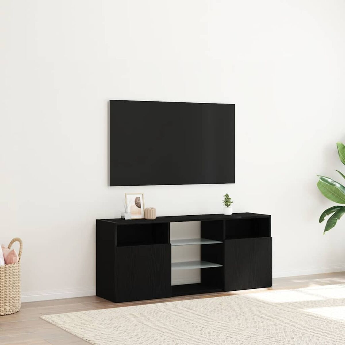 Ein schwarzer TV-Schrank mit einem Fernseher darüber. Auf dem Schrank stehen eine kleine Pflanze und ein gerahmtes Bild. Davor liegt ein Teppich.