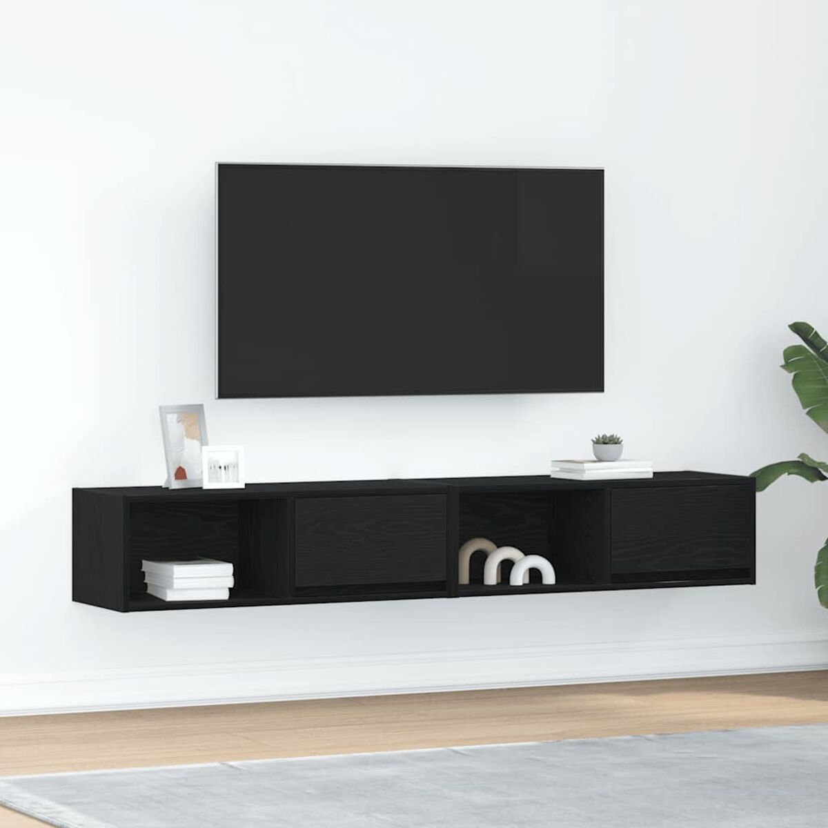 Ein schwarzer Fernseher ist über einem schwarzen, schwebenden Regal mit Dekoration angebracht. Rechts ist eine Pflanze zu sehen. Die Wand ist weiß.