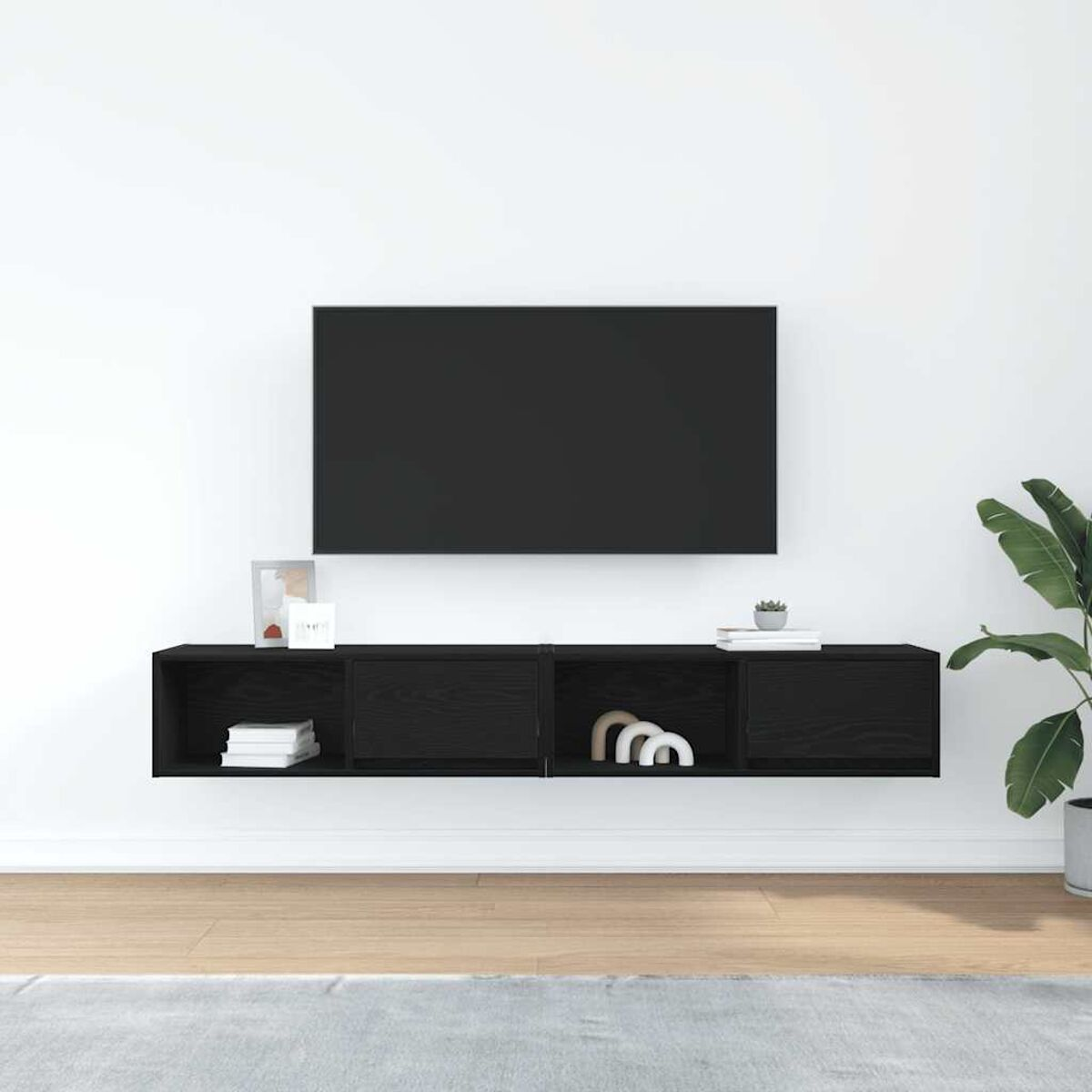 Ein schwarzer Fernseher hängt über einem schwarzen, an der Wand befestigten Schrank mit Dekoration. Rechts steht eine Pflanze. Holzfußboden und weiße Wand.