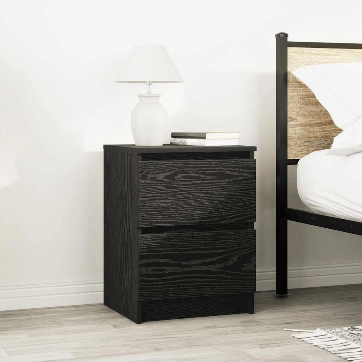 Schwarzer Nachttisch mit zwei Schubladen, einer Lampe und Büchern. Neben einem Bett mit weißem Kissen. Heller Holzboden und weiße Wand.