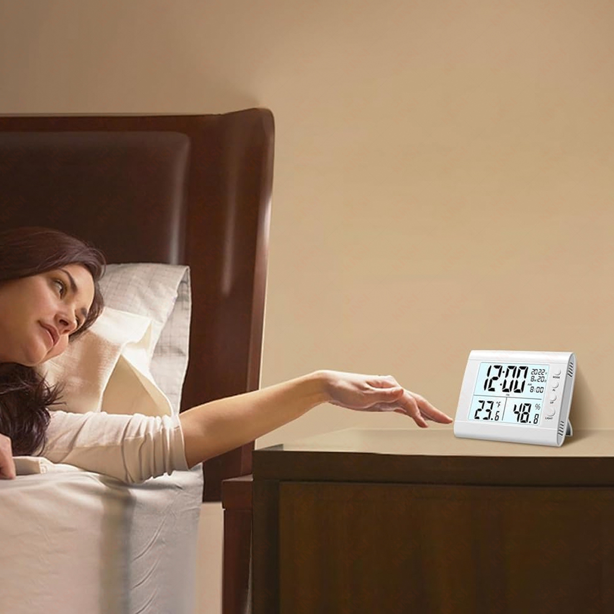 Frau im Bett greift nach einer Digitaluhr auf einem Nachttisch. Uhr zeigt Zeit, Temperatur und Luftfeuchtigkeit an.