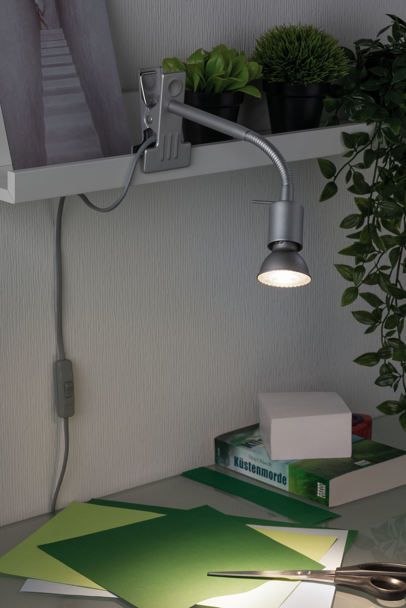 Lampa biurkowa oświetla zielony papier i nożyczki. Książka, biały klocek i rośliny znajdują się na półce powyżej.