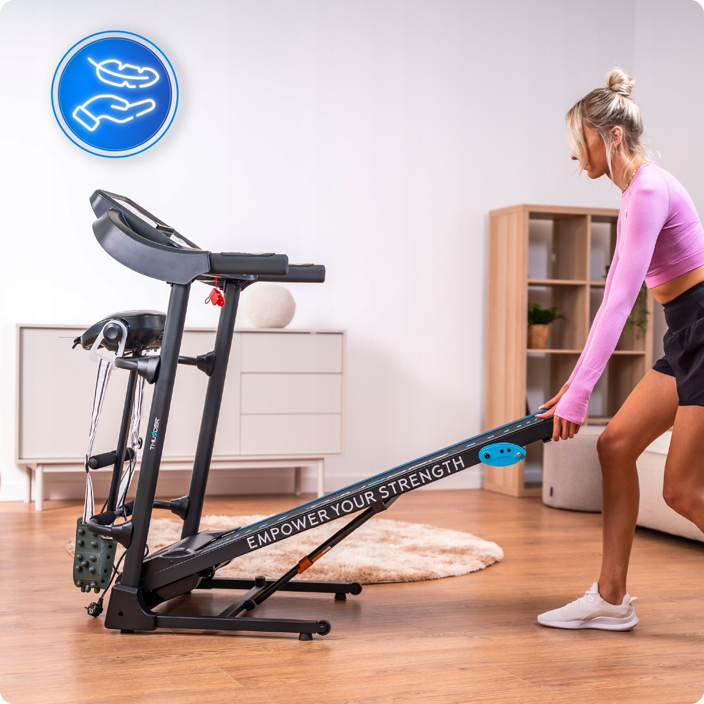 Kobieta w stroju do ćwiczeń opuszcza bieżnię. Bieżnia jest czarna z niebieskimi akcentami. Pokój jest jasny.