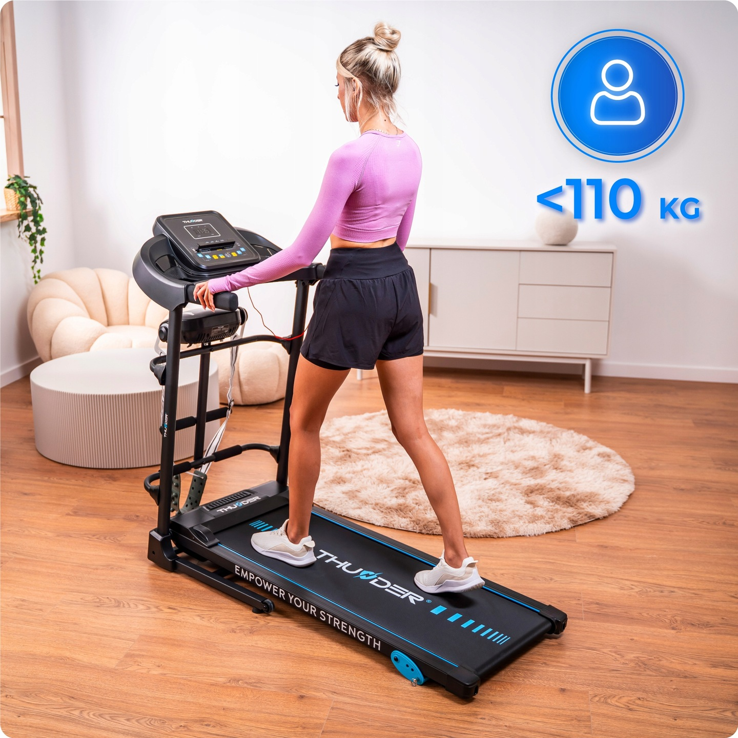 Kobieta w stroju sportowym stoi na bieżni. Bieżnia jest czarna i niebieska. Tekst na ścianie brzmi '<lt/>110 KG'.